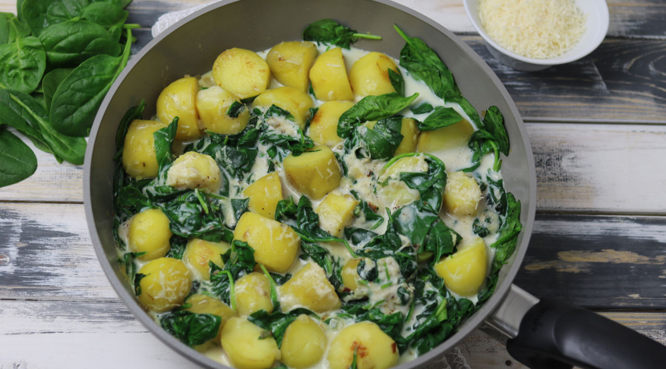 Eine Fissler-Pfanne mit Kartoffeln, Spinat und Rahmsauce auf Holz; frischer Spinat und geriebener Käse vervollständigen die erstklassige Szene.