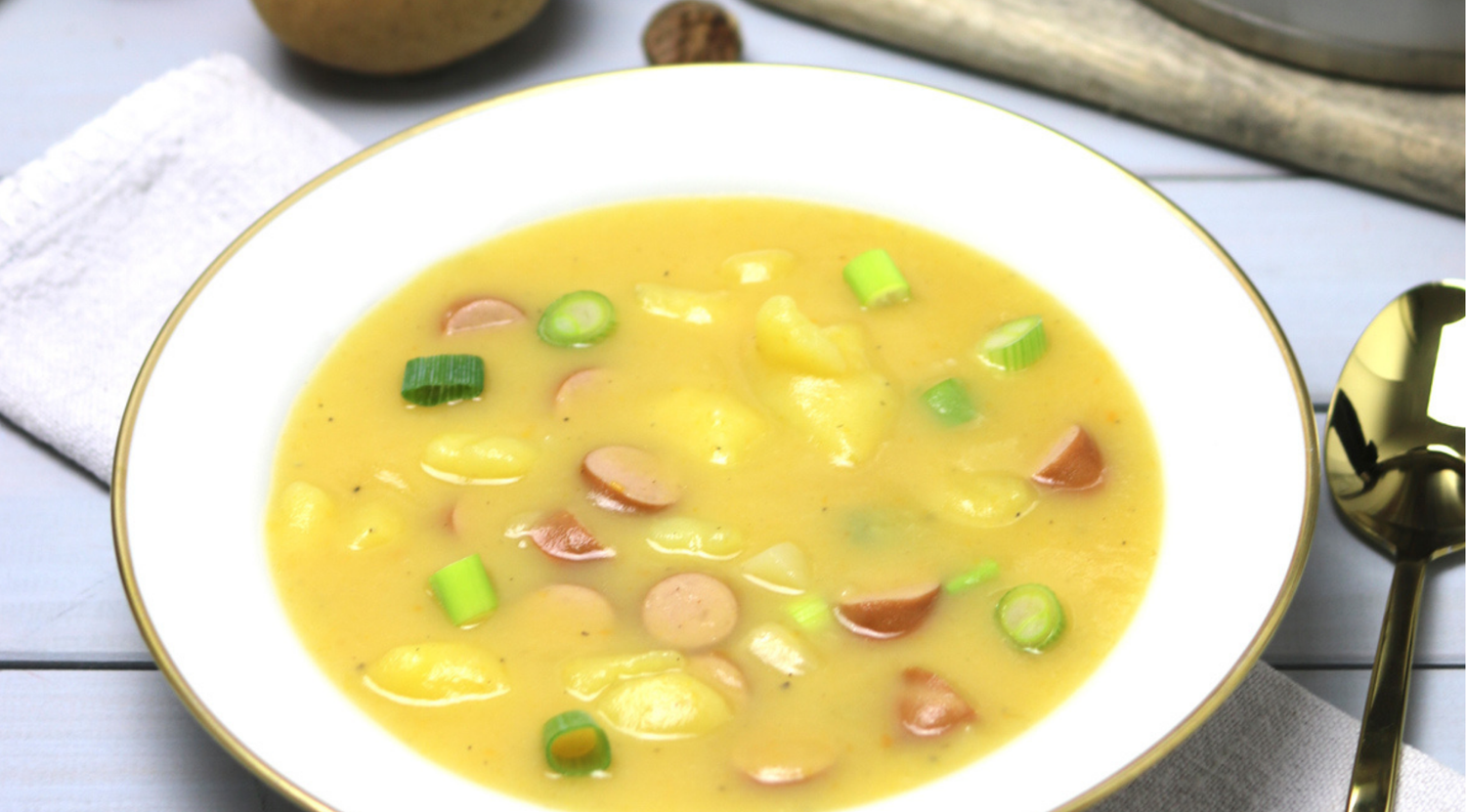 Kartoffelcremesuppe mit Würstchen und grünen Zwiebeln, elegant serviert nach Fissler-Art auf einem Teller mit Goldrand und Holztisch.