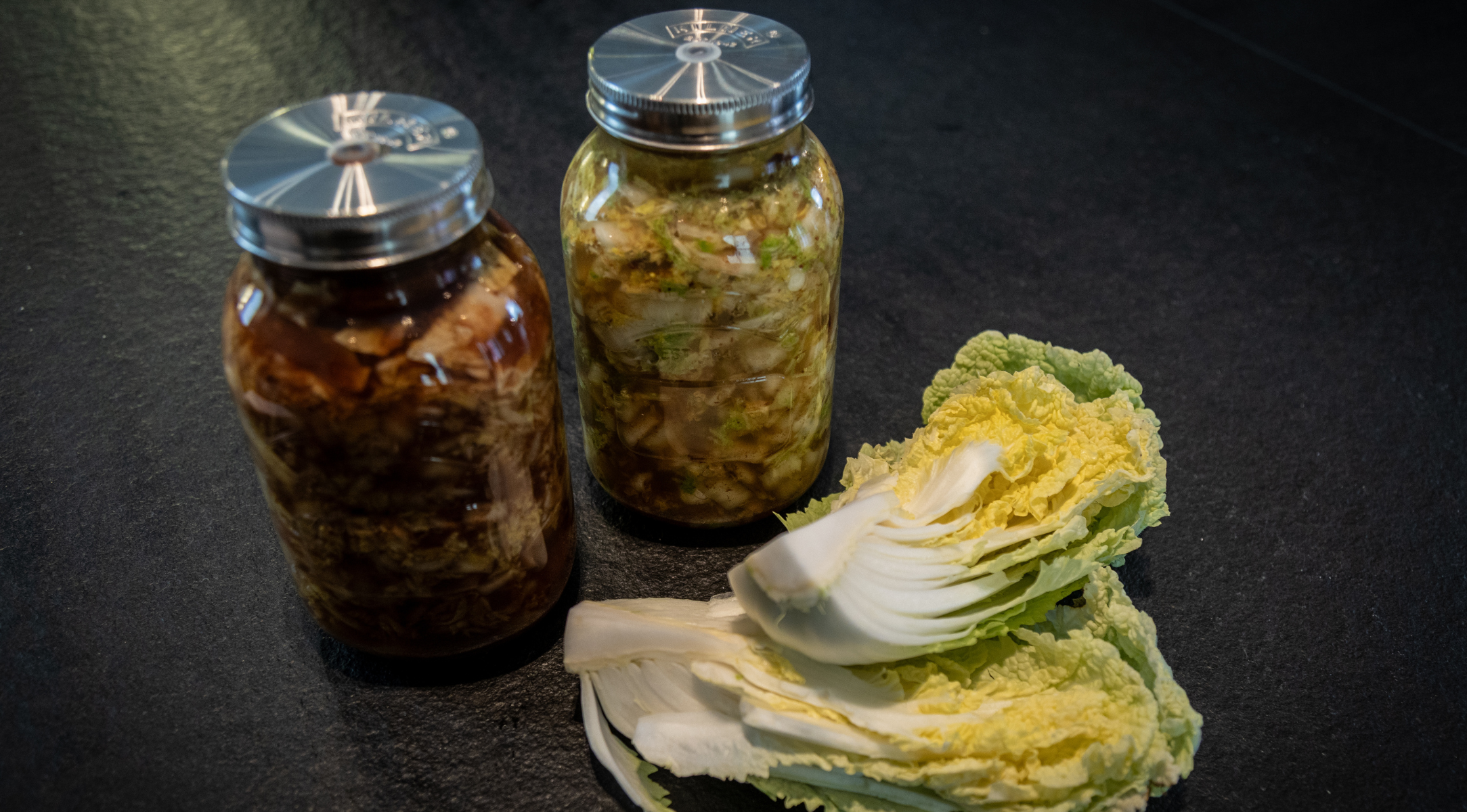 Zwei Gläser mit hausgemachtem Kimchi stehen auf einer dunklen Arbeitsplatte, flankiert von frischen Chinakohlblättern – einige ganz, andere halbiert –, und schaffen so eine einladende Küchenszene. Diese mit Präzision und Sorgfalt gestaltete Kulisse unterstreicht die Kunst der Fermentierung, die mit dem langlebigen Kochgeschirr von Fissler ganz einfach wird – entworfen in Deutschland für zeitlose Qualität und Innovation.