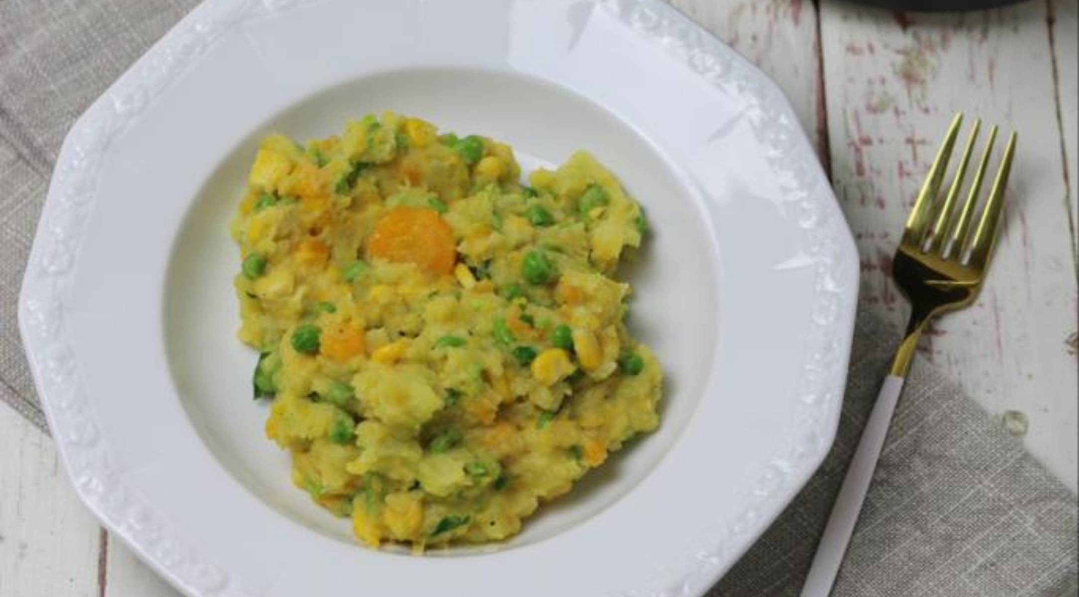 Gemüsepüree - Erbsen, Mais, Karotten, Kartoffeln - auf einem weißen Teller mit einer goldenen Gabel, auf einem Holztisch mit Serviette.