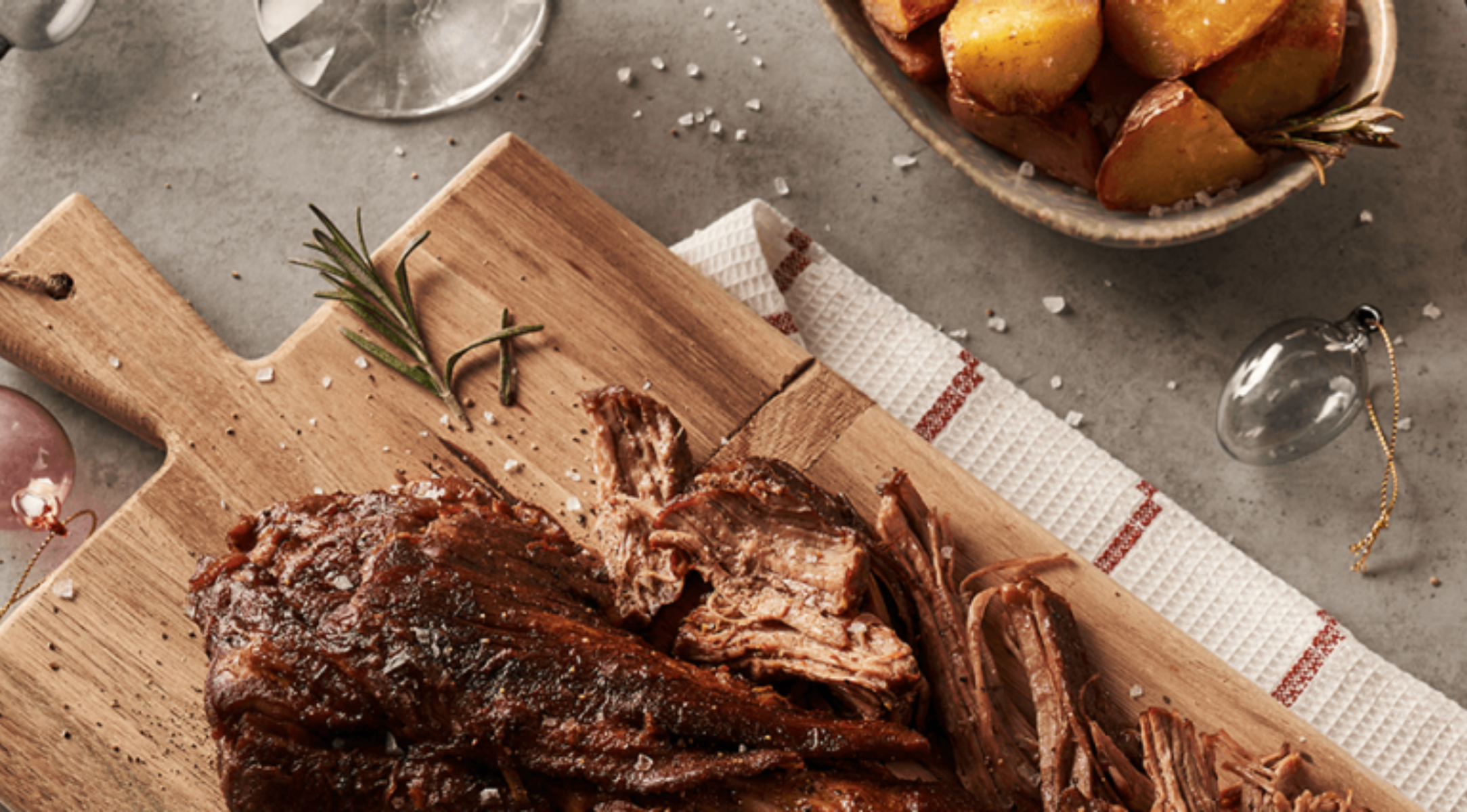 Zerkleinertes Bratenfleisch mit Rosmarin auf einem Holzbrett, dazu Röstkartoffeln, serviert mit Fissler-Gläsern und Küchenzubehör.