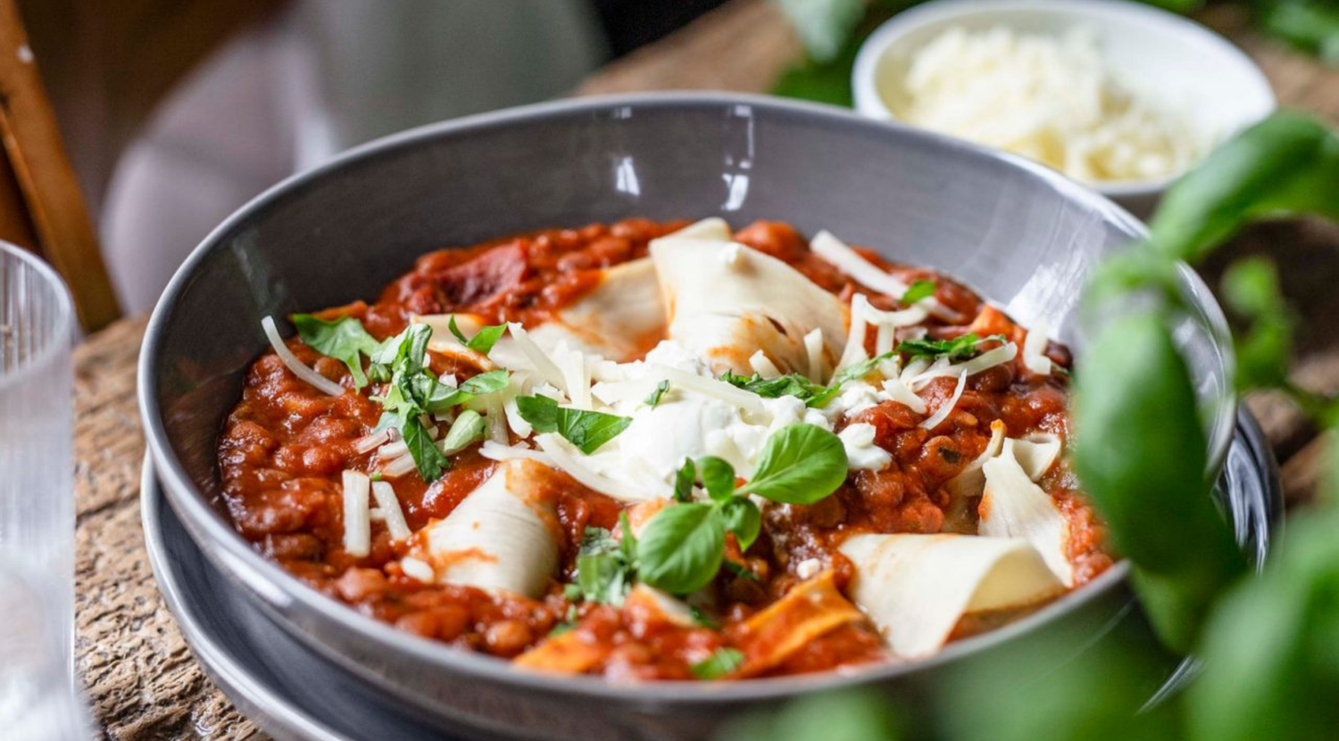 Lasagne-Suppe in einer hochwertigen Fissler-Schale auf einem rustikalen Tisch serviert, mit Basilikum und Käse überbacken. Daneben eine Schale mit geriebenem Käse.