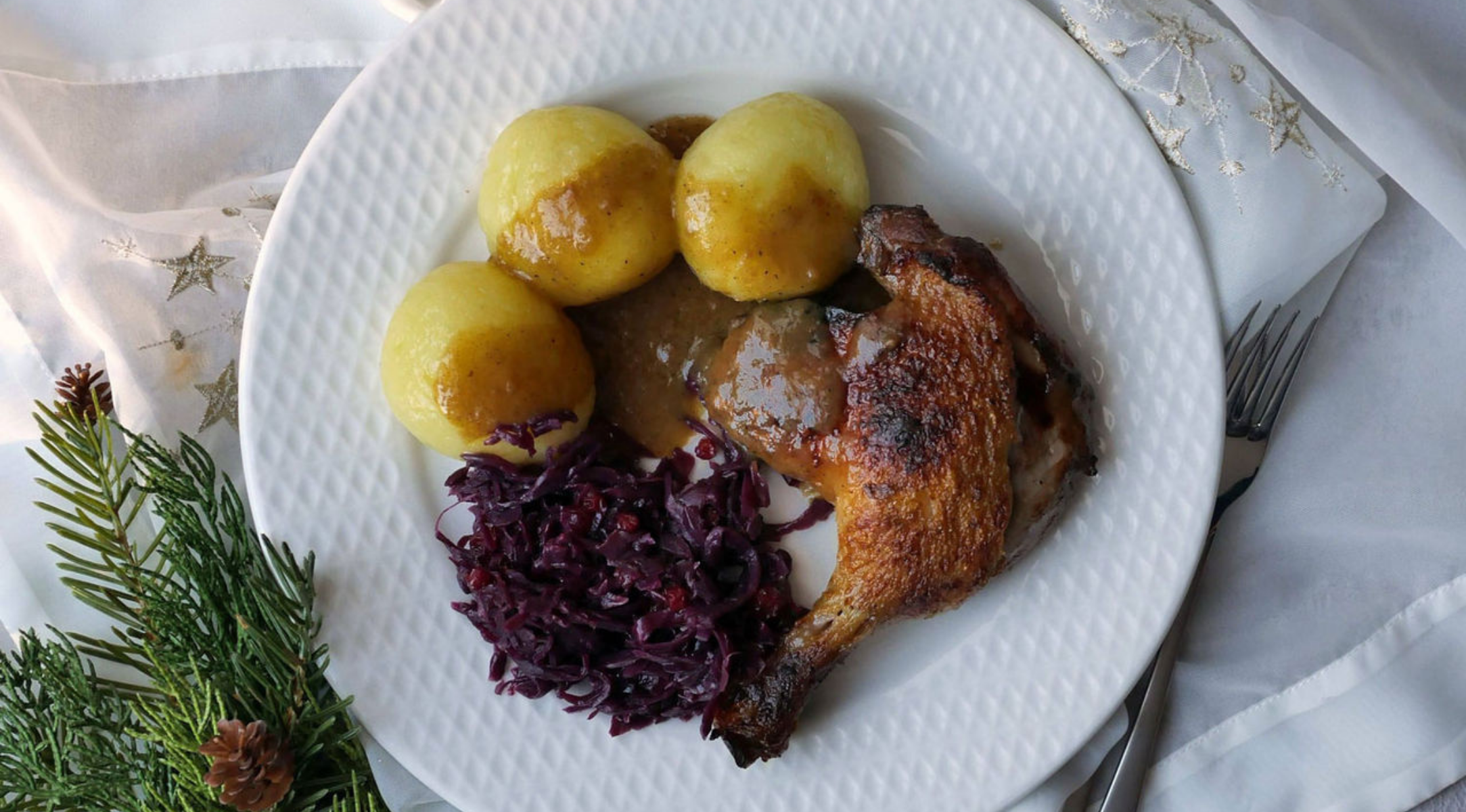 Ein festlicher Teller mit Entenkeule, Kartoffelklößen und Rotkohl, stilisiert neben Kiefer auf einem weißen Tuch - perfekt aus Fissler-Kochgeschirr.