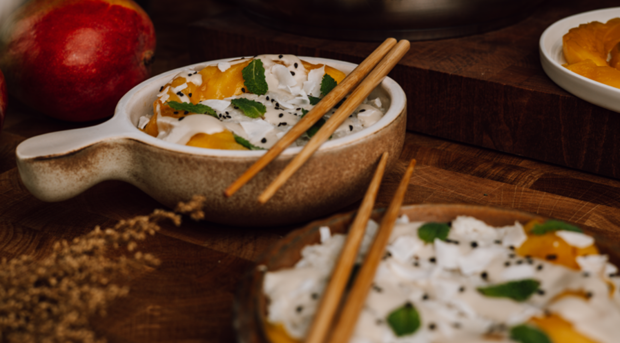 Eine Keramikschüssel mit geschnittener Mango, Kokosnusscreme und Minze wird mit Fissler-Stäbchen serviert; im Hintergrund ist eine ganze Mango zu sehen.