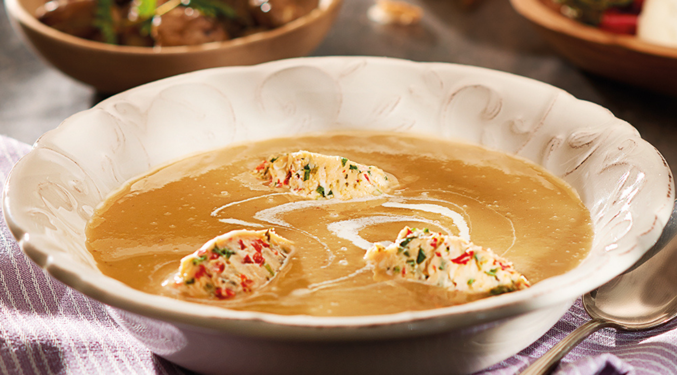 Eine Schüssel mit brauner Cremesuppe mit Kräuterkäse, elegant serviert auf einem gestreiften Tuch neben einem Fissler-Edelstahllöffel.