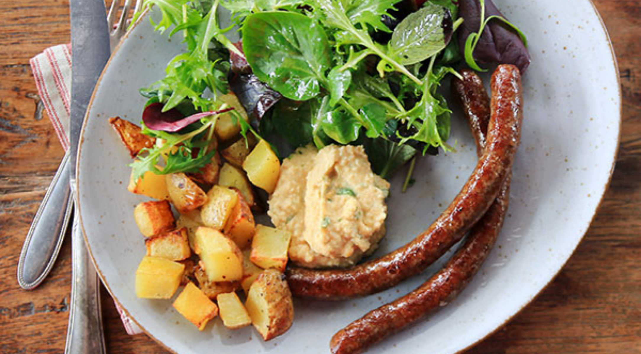 Eine Mahlzeit mit Würstchen, gebratenen Kartoffeln, Hummus und Salat, serviert auf einem Holztisch, mit Besteck daneben - ein Präzisionskochgeschirr.