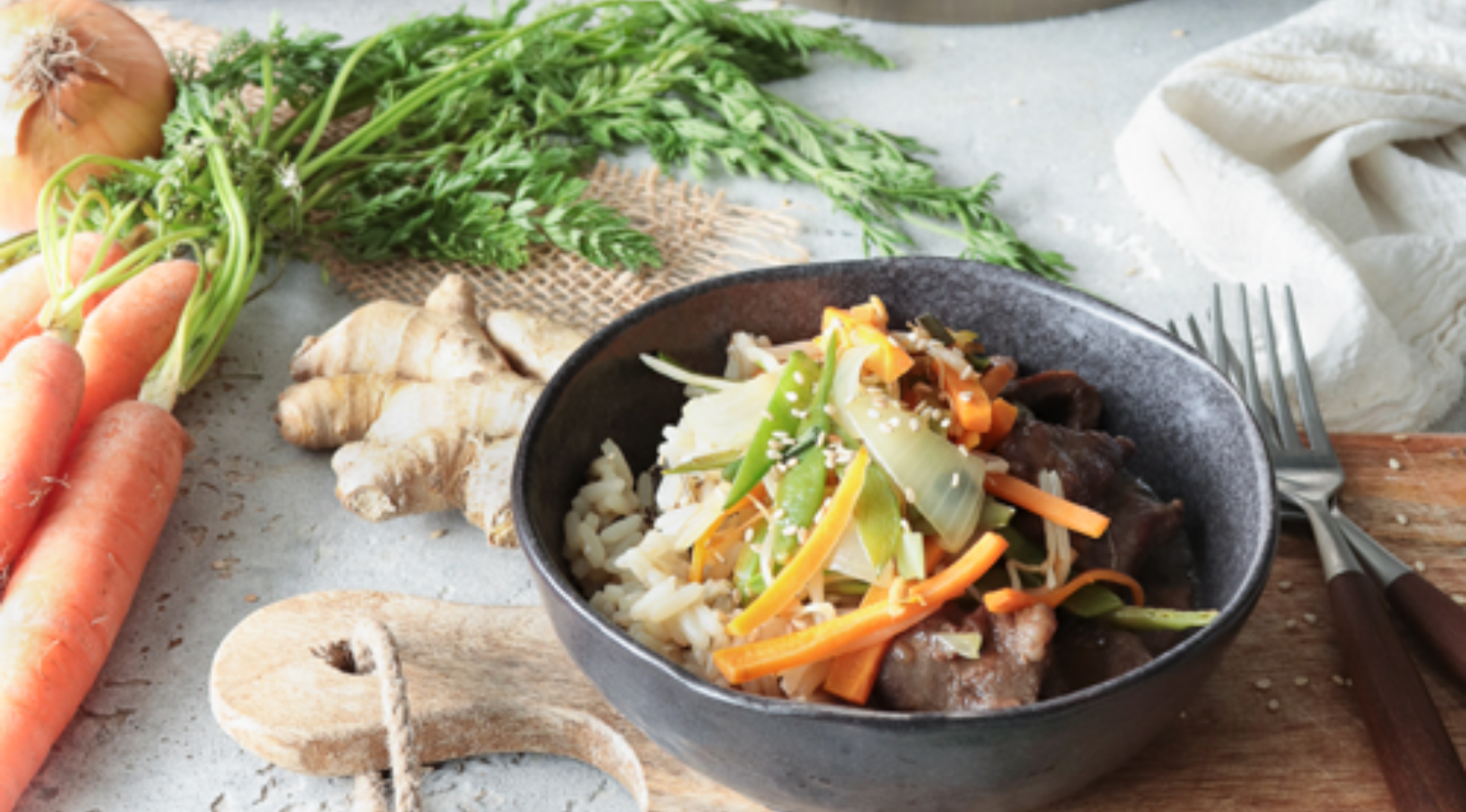 Reisschüssel mit gebratenem Rindfleisch, Gemüse und Sesam, daneben frisches Gemüse. Kochgeschirr und Besteck von Fissler vervollständigen die Szene.