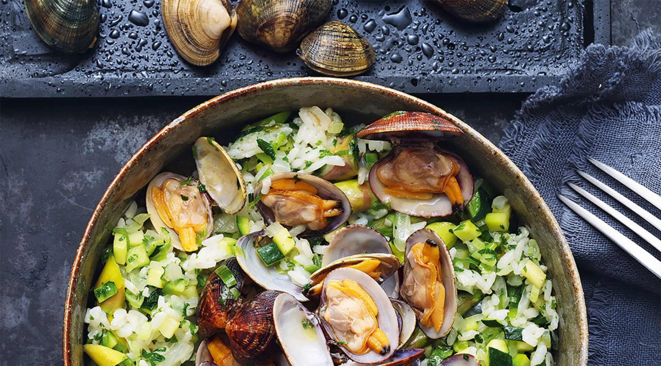 Eine lebendige Schale Reis mit Venusmuscheln neben frischen Venusmuscheln, dargestellt mit dem Premium-Besteck von Fissler auf einer eleganten dunklen Oberfläche.
