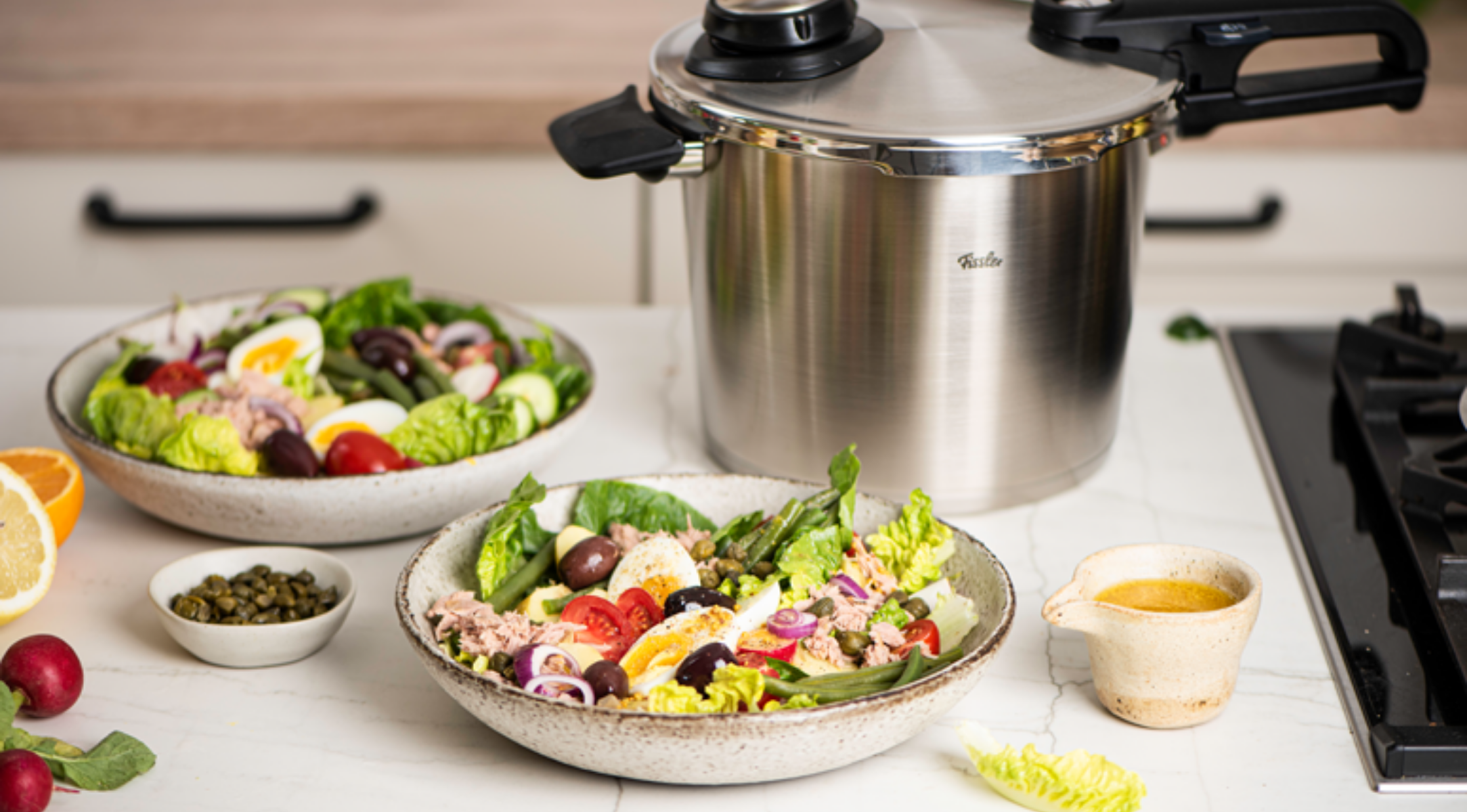 Ein Fissler Schnellkochtopf aus rostfreiem Stahl auf einer Küchentheke mit frischen Salaten, Kapern, Soßenkrügen und Gemüse.