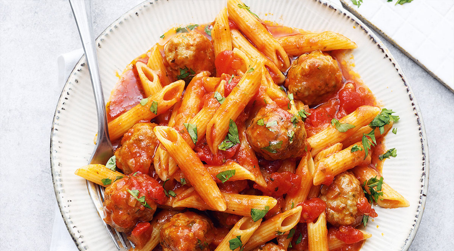 Penne mit Tomatensoße, Fleischbällchen und Petersilie, serviert auf einem makellosen weißen Teller, der mit Fissler-Qualitätskochgeschirr hergestellt wurde.