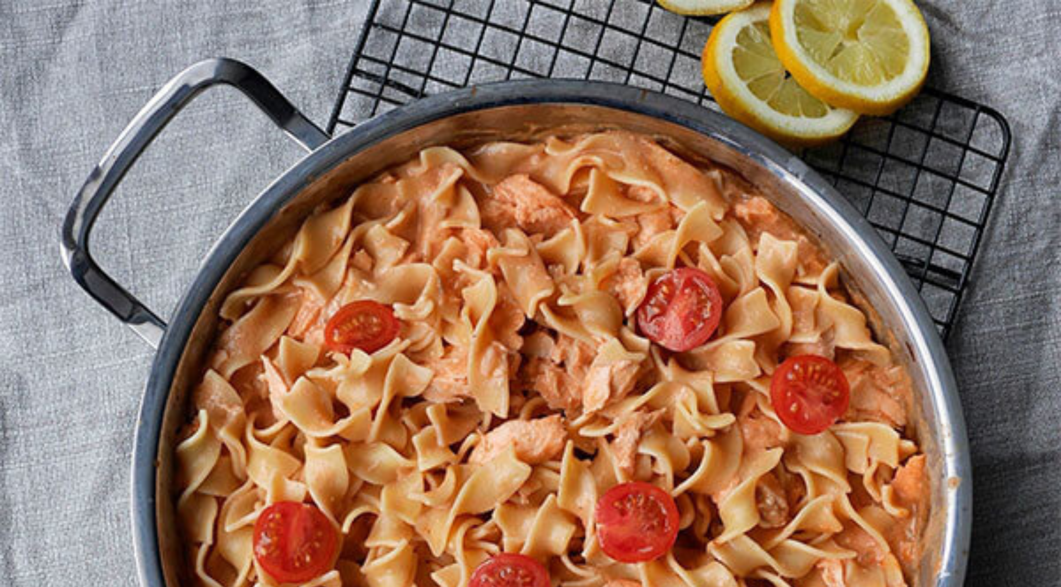 Eine Fissler-Edelstahlpfanne mit cremiger Lachspasta und Tomaten auf einem Rost, daneben Zitronenscheiben - Präzision trifft auf zeitloses Design.