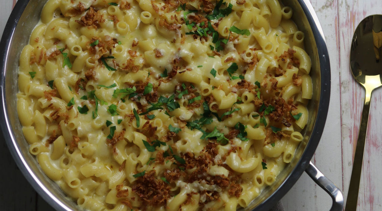 Fissler-Premium-Pfanne mit Makkaroni und Käse, knusprigen Zwiebeln, Petersilie garniert; Goldlöffel daneben auf einer weißen Holzunterlage.