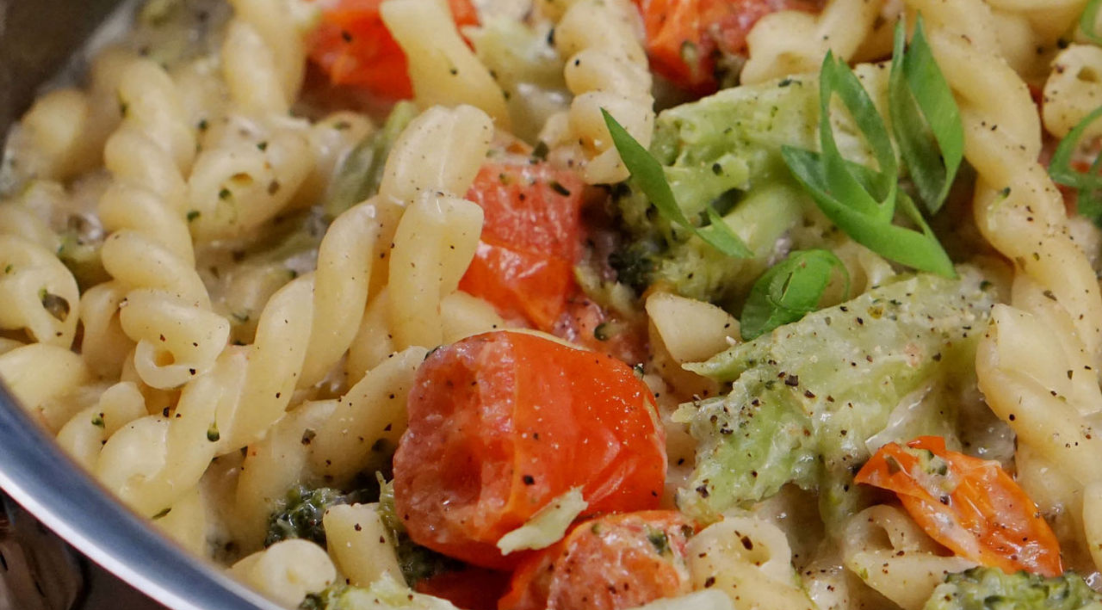 Cremige Nudelspiralen, geröstete Kirschtomaten, Brokkoli und Frühlingszwiebeln, zubereitet in einer Fissler-Edelstahlpfanne.