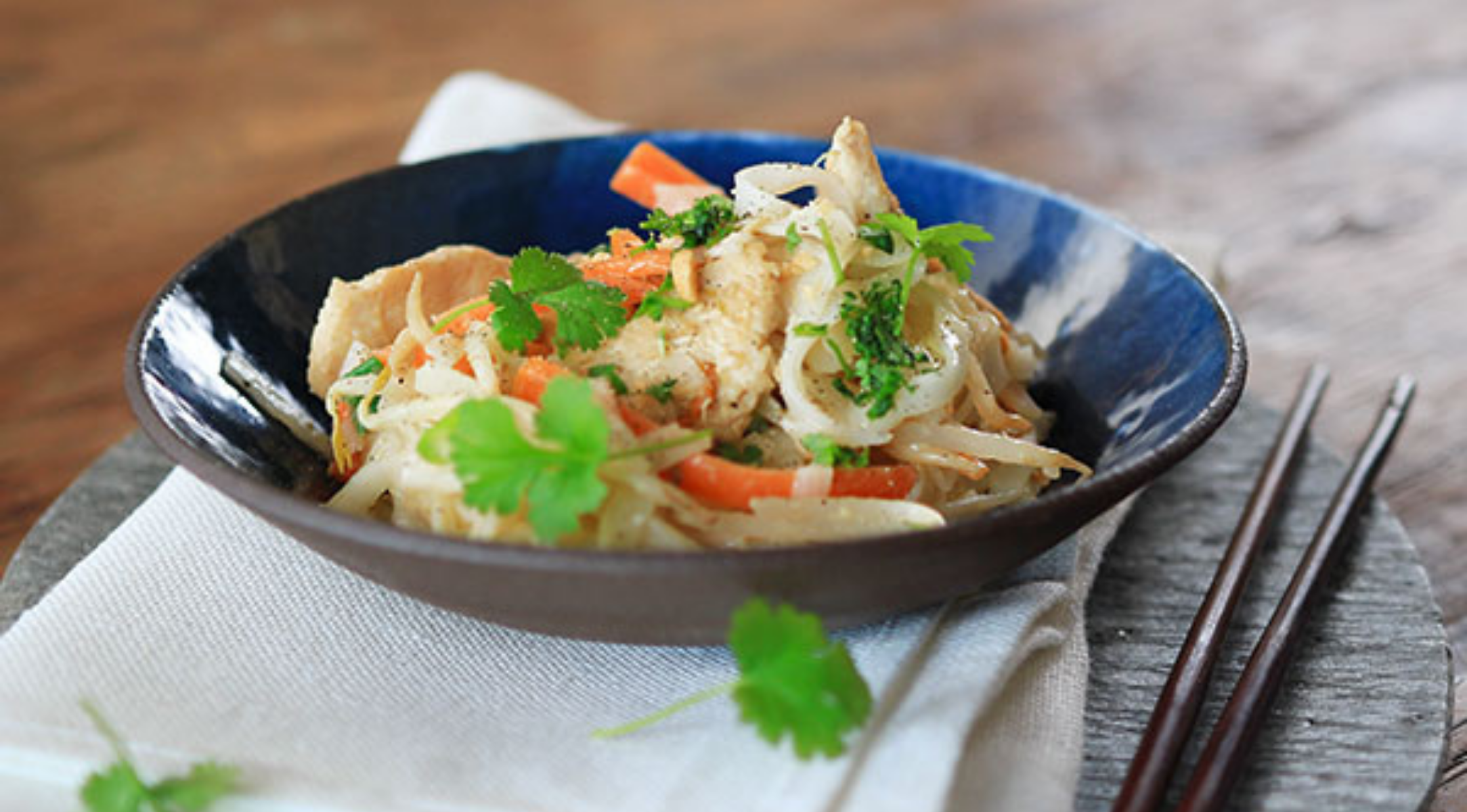 Asiatisches Nudelpfannengericht mit Hühnchen, Karotten und Brokkoli in einer Schüssel - exquisit präsentiert auf einem von Fissler gefertigten Holztischset.