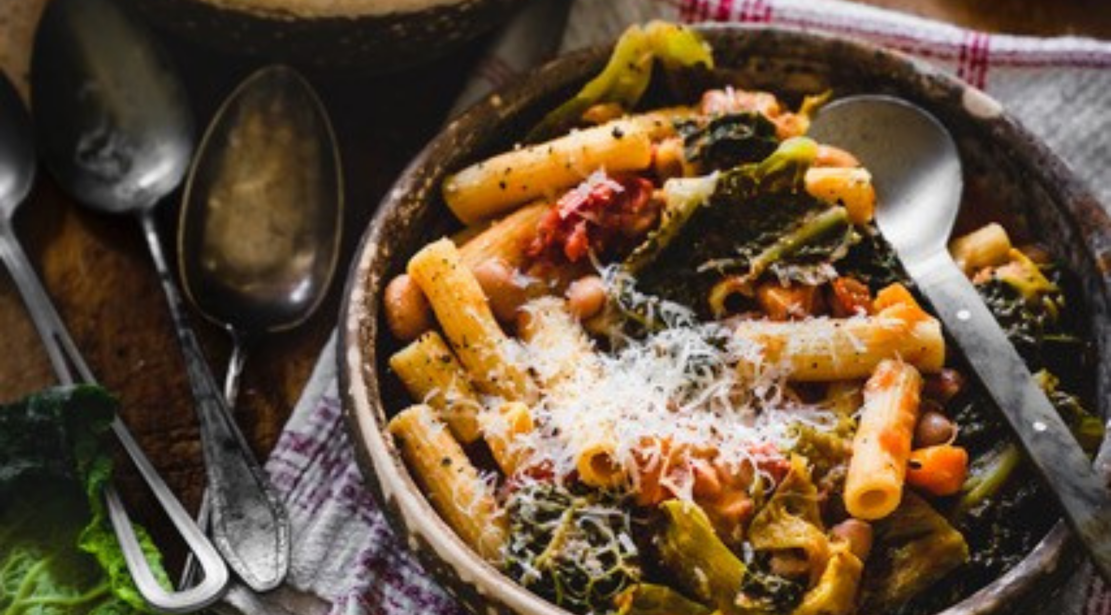 Eine Schüssel mit Rigatoni, belegt mit Gemüse, Bohnen, Tomatensauce und Käse. Das Fissler-Besteck liegt auf einer gestreiften Serviette daneben.