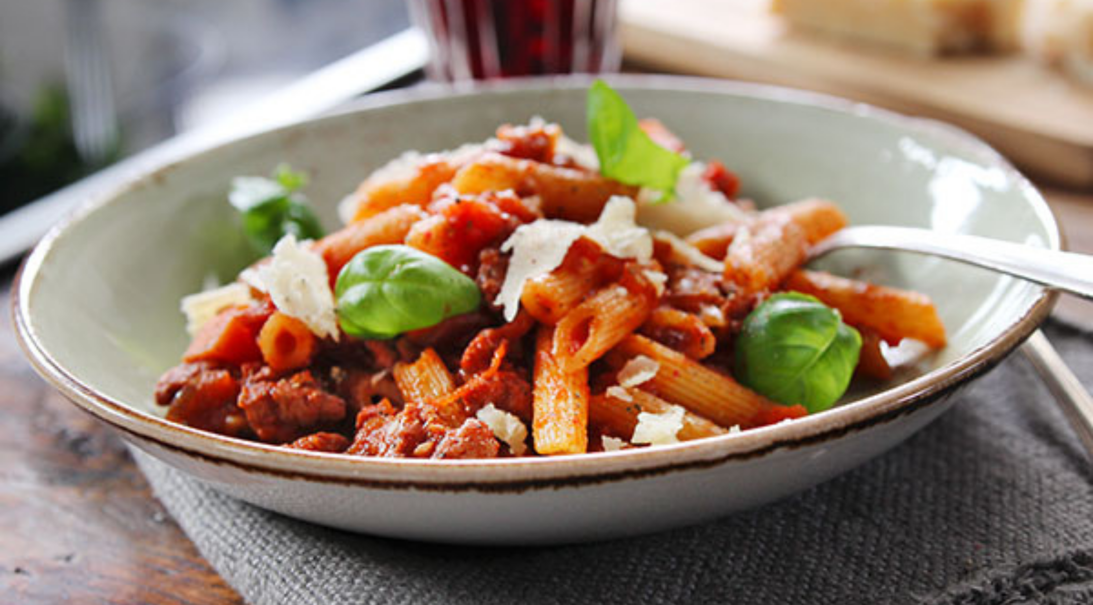Penne-Nudeln mit Tomatensauce, Hackfleisch, Basilikum und Käse auf einem rustikalen Teller, serviert mit einer Gabel - hergestellt in Fissler-Kochgeschirr.