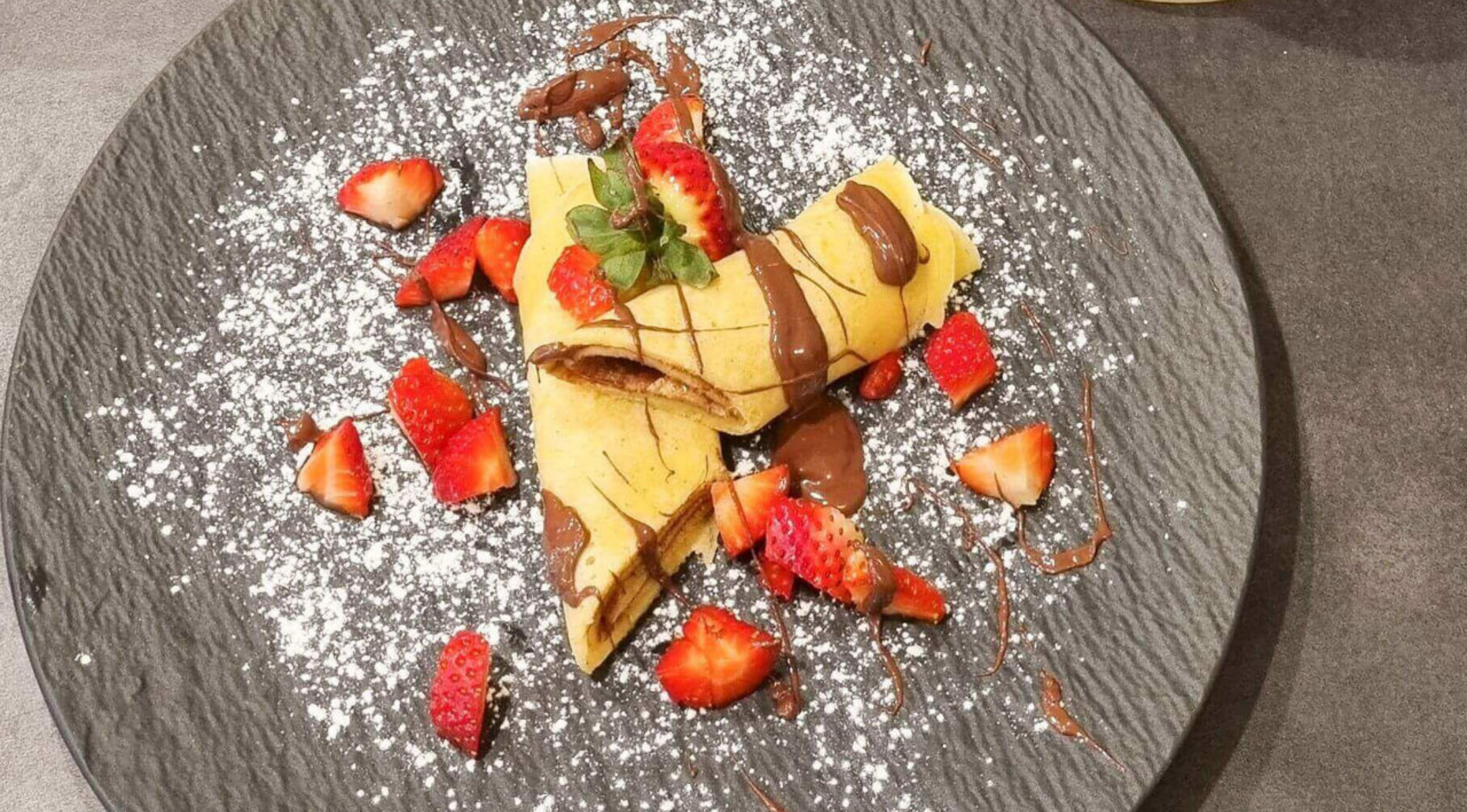 Zwei gerollte Crêpes mit Schokostreuseln, Erdbeeren und Minze auf einem dunklen Teller, elegant präsentiert auf hochwertigem Fissler-Geschirr.