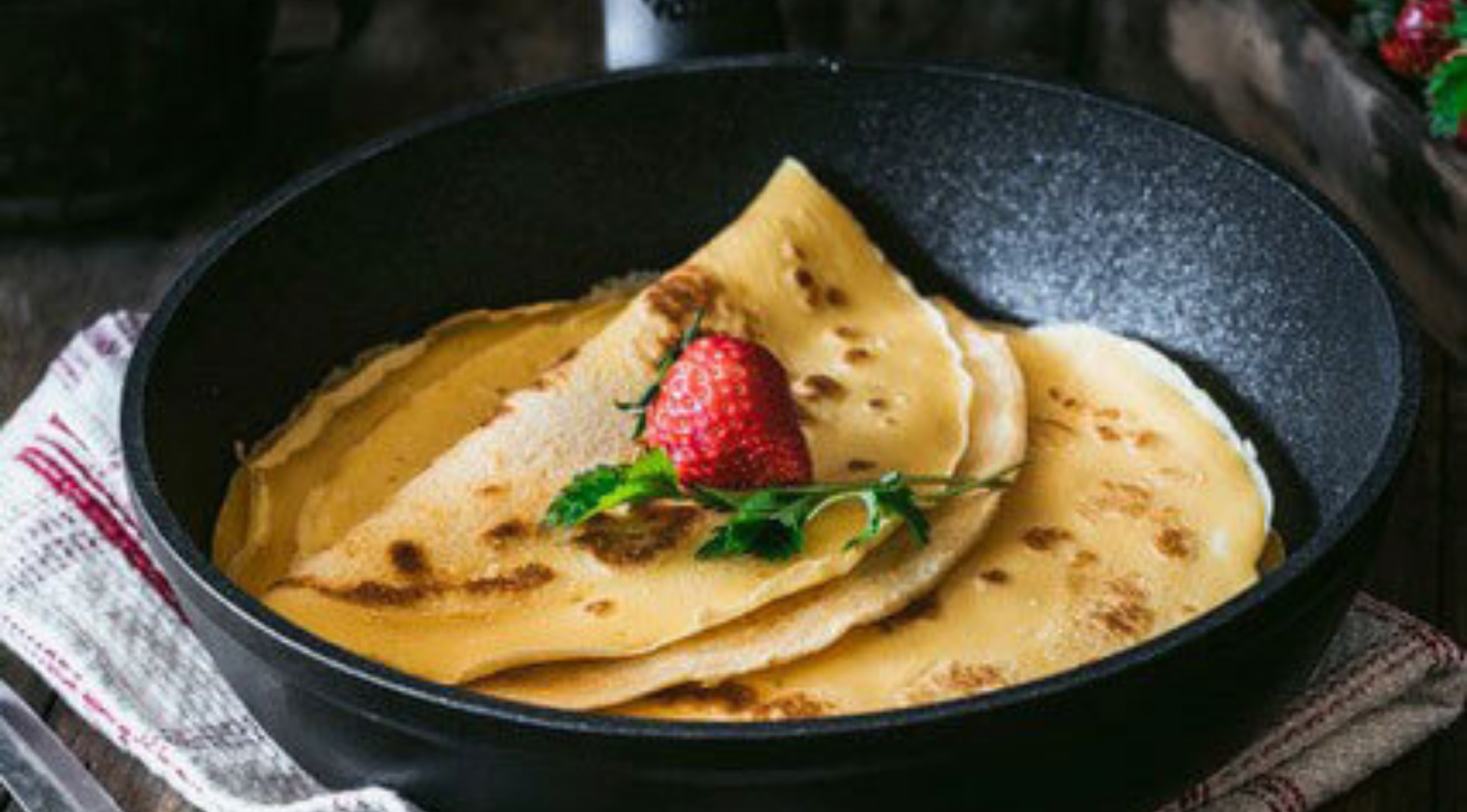 Zwei Crêpes mit Erdbeeren und Petersilie ruhen in einer eleganten Fissler-Pfanne, die deutsche Handwerkskunst auf einem gestreiften Küchentuch präsentiert.