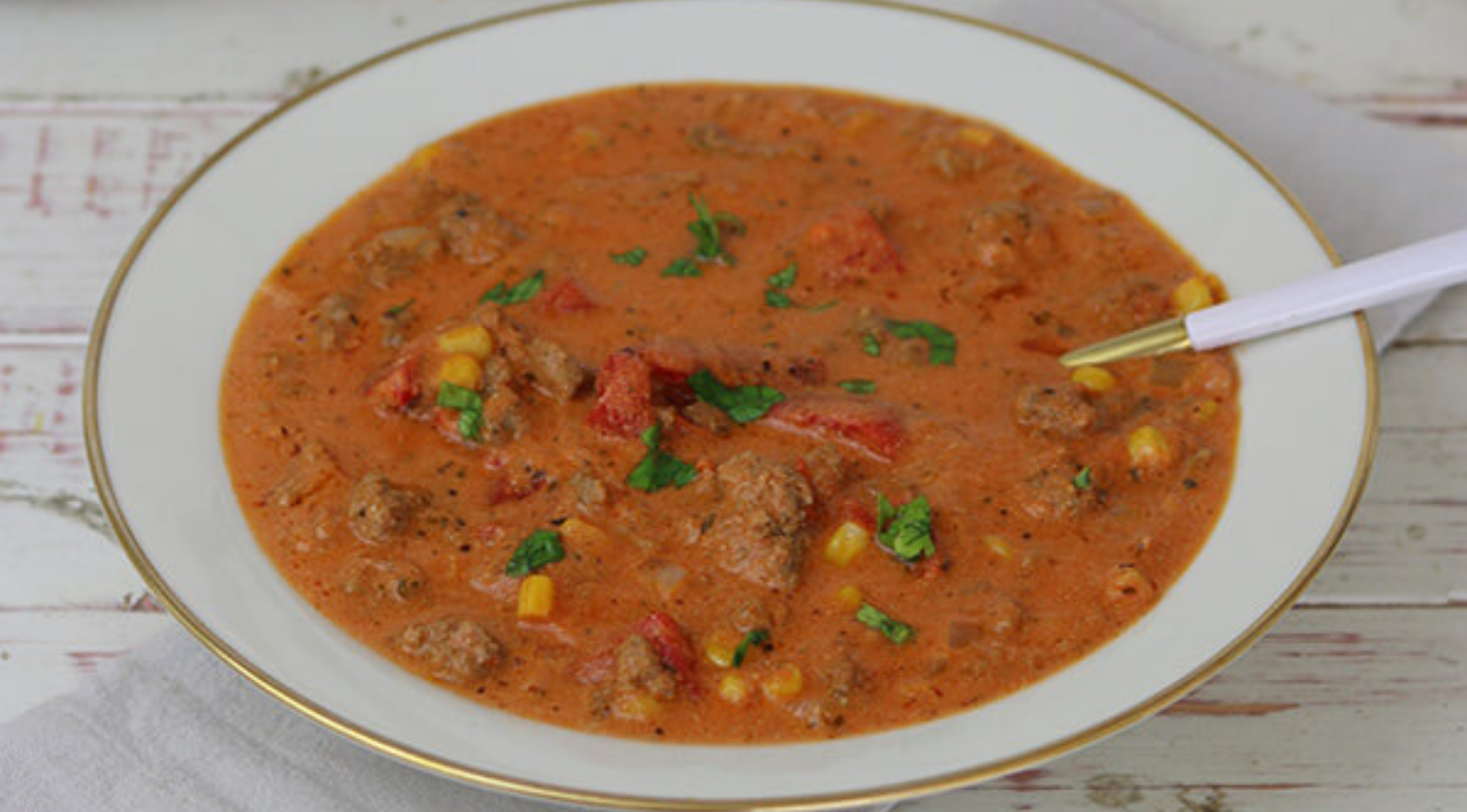 Eine weiße Schale mit herzhafter Tomatensuppe und Kräutern steht auf einem weißen Tisch, dazu ein Löffel - perfekt zubereitet im Fissler-Kochgeschirr.