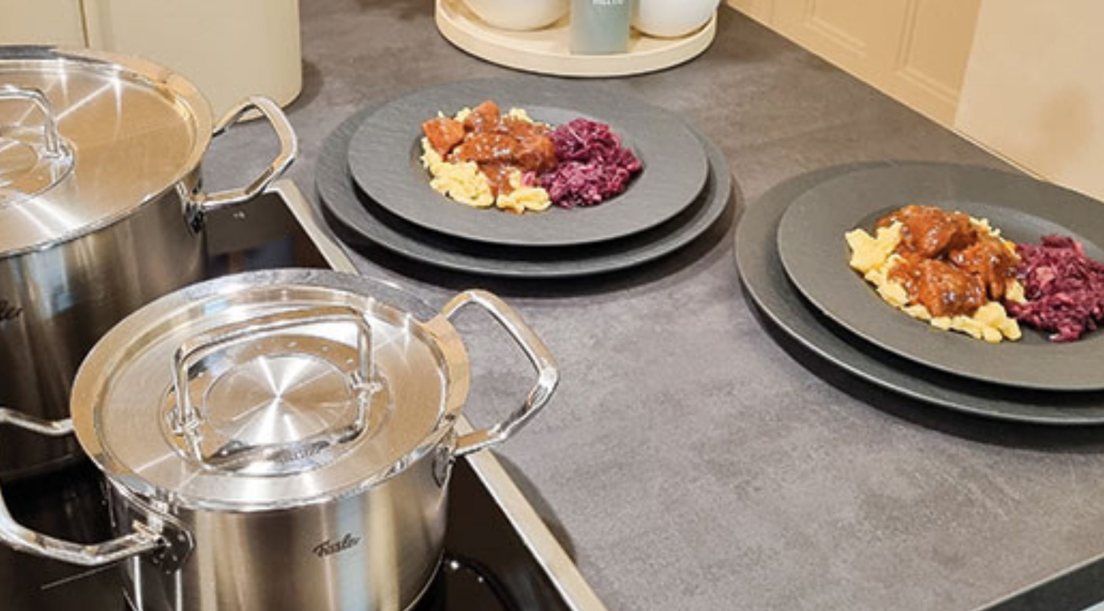 Zwei Fissler-Edelstahltöpfe mit Deckeln stehen auf einem Herd neben Tellern mit Eintopf, lila Kraut und Nudeln auf einem Küchentisch.