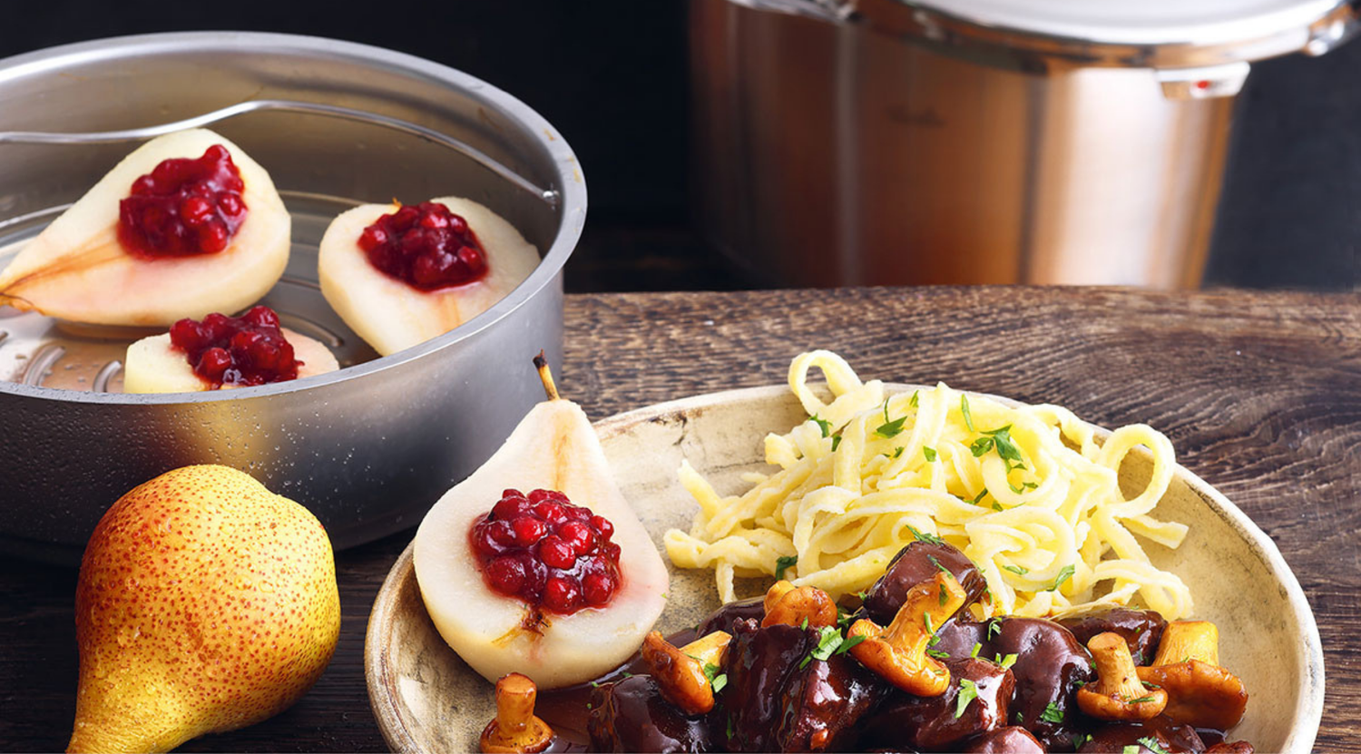 Nudeln, Pilzeintopf und mit Beeren gefüllte Birnen neben einer frischen Birne, Fissler-Edelstahlkochgeschirr auf Holzoberfläche.