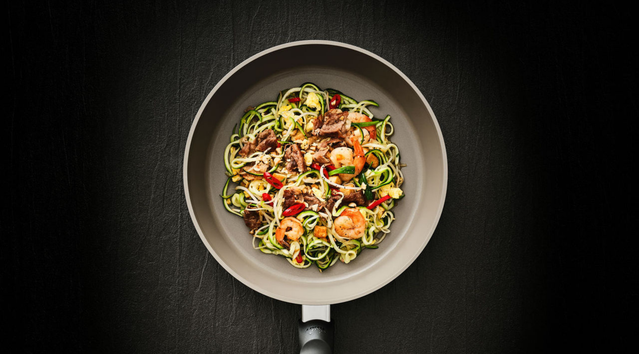 Eine Fissler-Pfanne auf dunkler Oberfläche enthält Zucchini-Nudeln, Garnelen, Rindfleisch, Chili, grüne Zwiebeln und Rührei - alles in Präzisionsarbeit hergestellt.