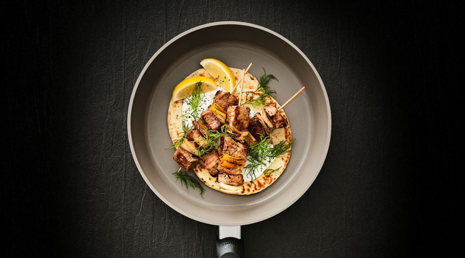 Eine Fissler Bratpfanne mit gegrillten Fleischspießen, Fladenbrot, Zitronenspalten und Kräutern, die deutsches Präzisionskochgeschirrdesign demonstriert.
