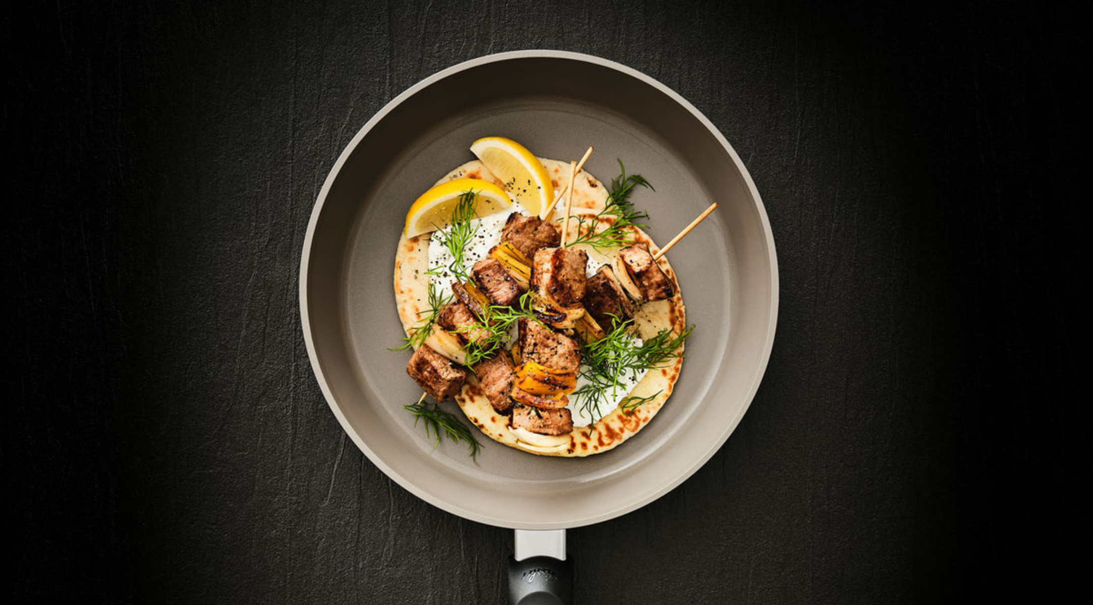 Eine Fissler Bratpfanne mit gegrillten Fleischspießen, Fladenbrot, Zitronenspalten und Kräutern, die deutsches Präzisionskochgeschirrdesign demonstriert.