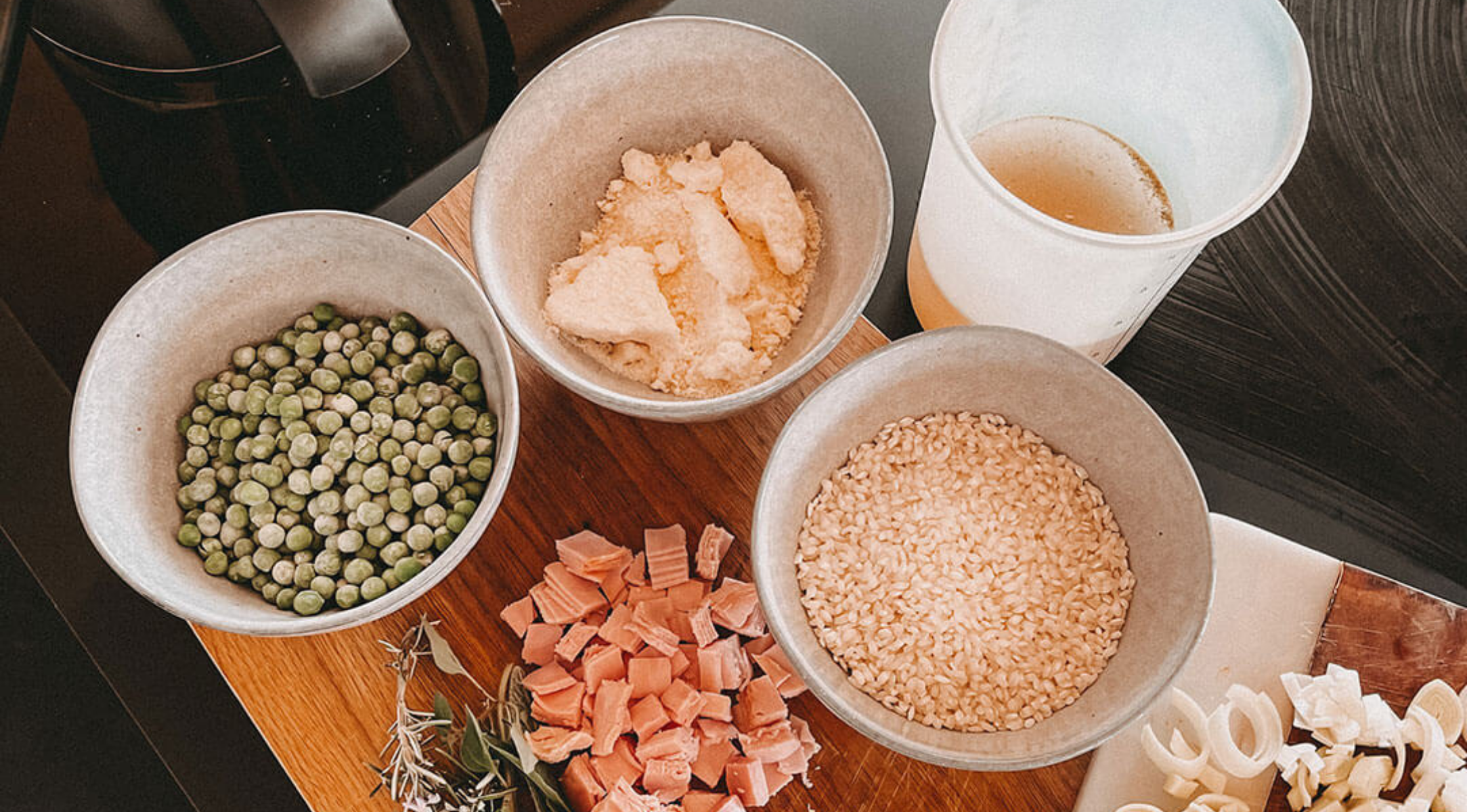 Auf einer Fissler-Tafel stehen Erbsen, Käse, Reis, Schinken, Brühe und frische Kräuter - Zutaten, die für hochwertige deutsche Kochtöpfe geeignet sind.