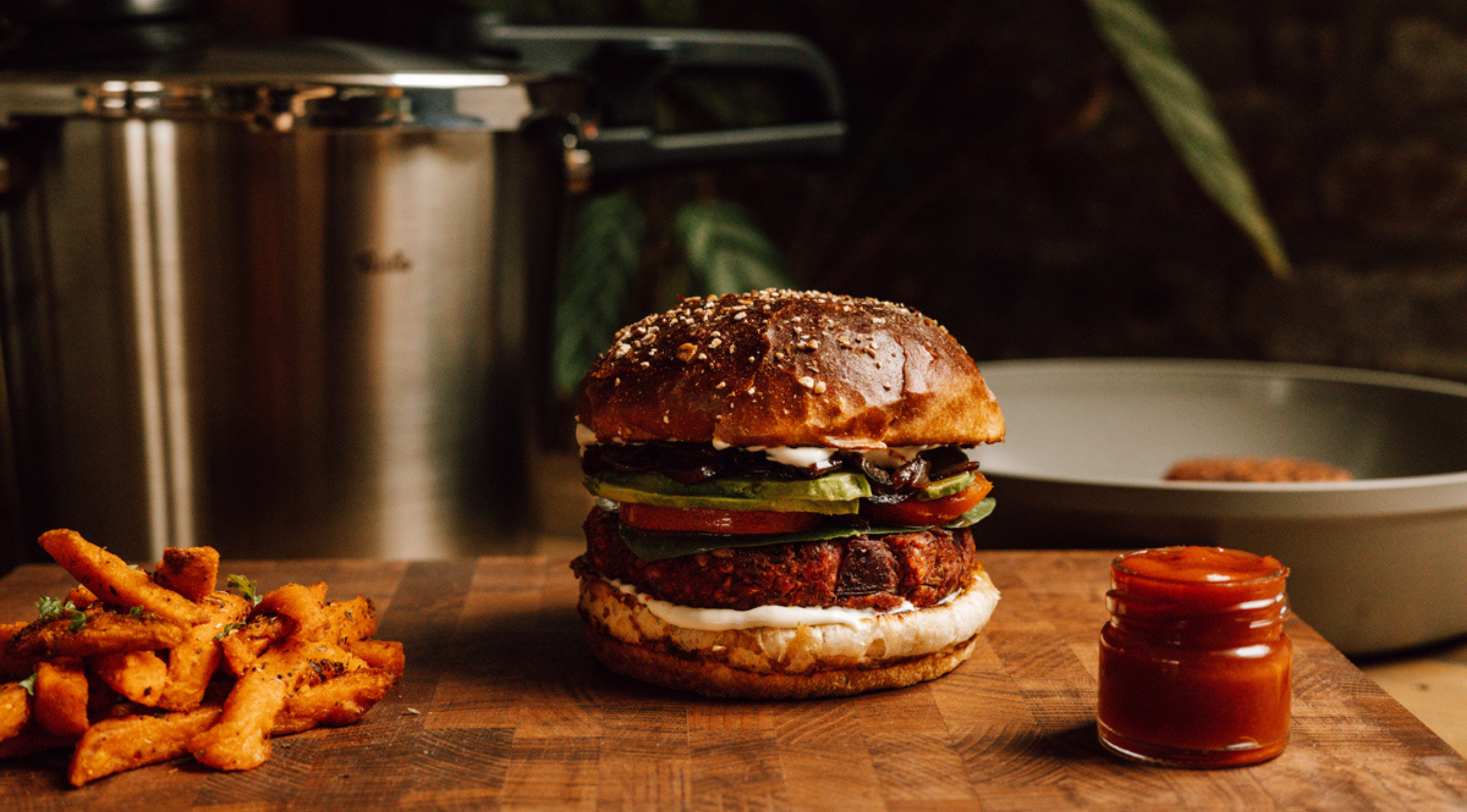 Ein Veggie-Burger mit Beilagen auf einem Holzbrett; im Hintergrund sorgen ein hochwertiger Fissler-Edelstahltopf und eine Schüssel für Eleganz.