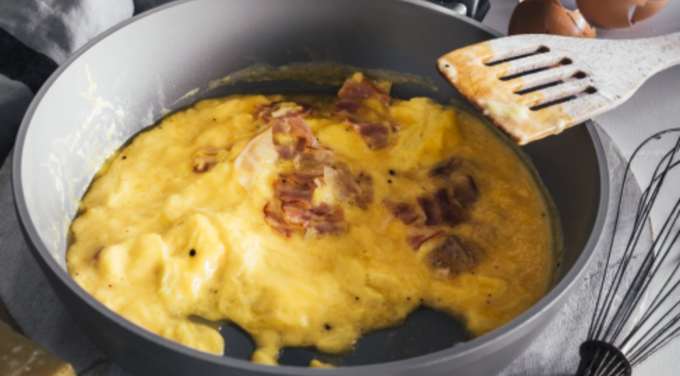 Rührei und Schinken kochen in einer Fissler-Pfanne neben einem Holzspatel, Eierschalen und einem Schneebesen auf einer glatten Arbeitsplatte.