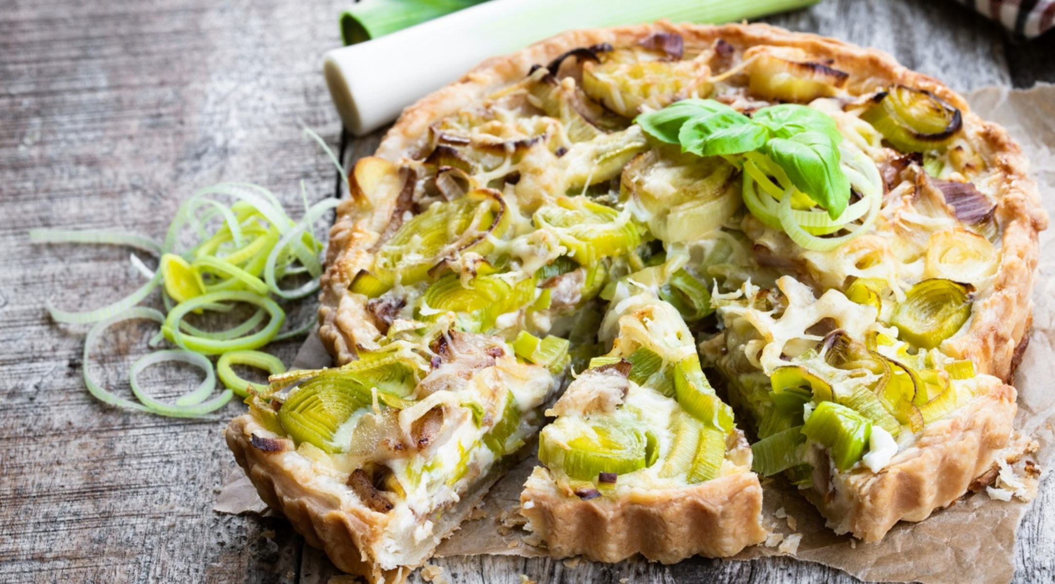 Eine goldene Lauch-Quiche, in Scheiben geschnitten und garniert, ruht auf Holz. Frischer Lauch, der die zeitlose Qualität des Fissler Kochgeschirrs unterstreicht.