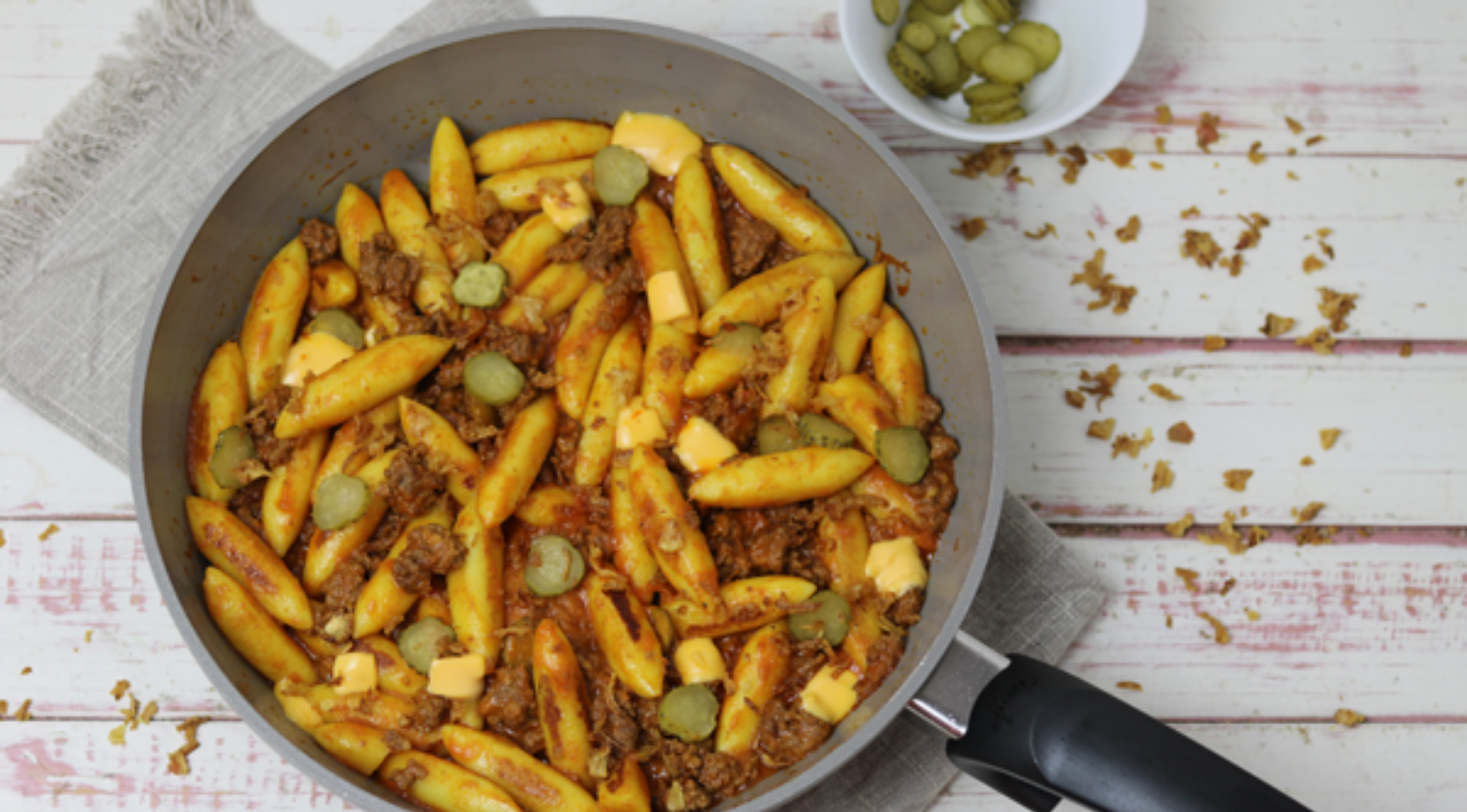 Eine hochwertige Fissler-Pfanne mit Penne, Hackfleisch, Käse und Essiggurken auf einem rustikalen Tisch; zusätzliche Essiggurken in einer Schale daneben.