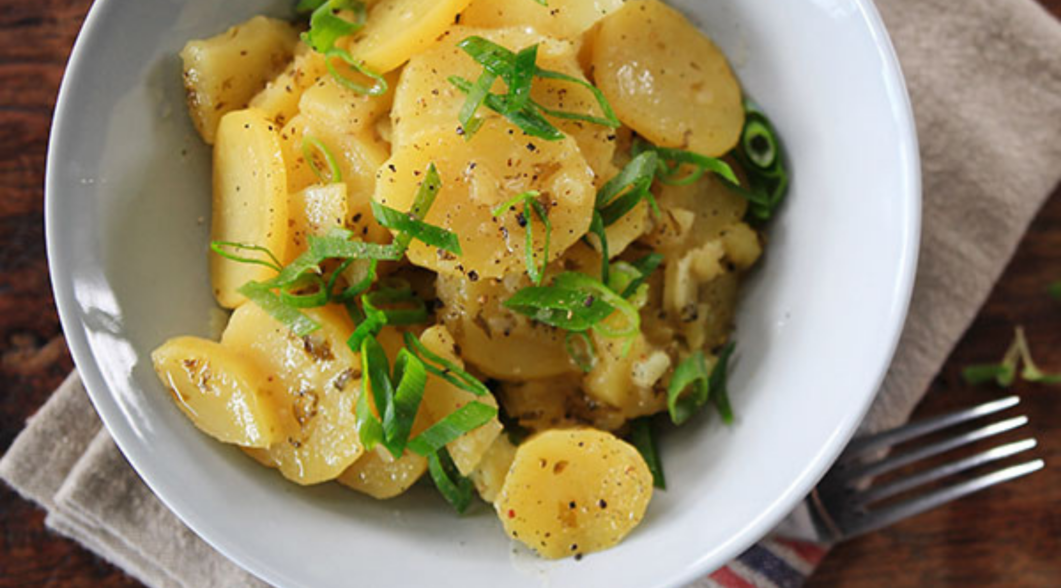 In Scheiben geschnittene Kartoffeln mit grünen Zwiebeln und schwarzem Pfeffer in einer weißen Schüssel auf beigefarbenem Stoff - präzise plattiert, von Fissler-Kochgeschirr inspiriert.