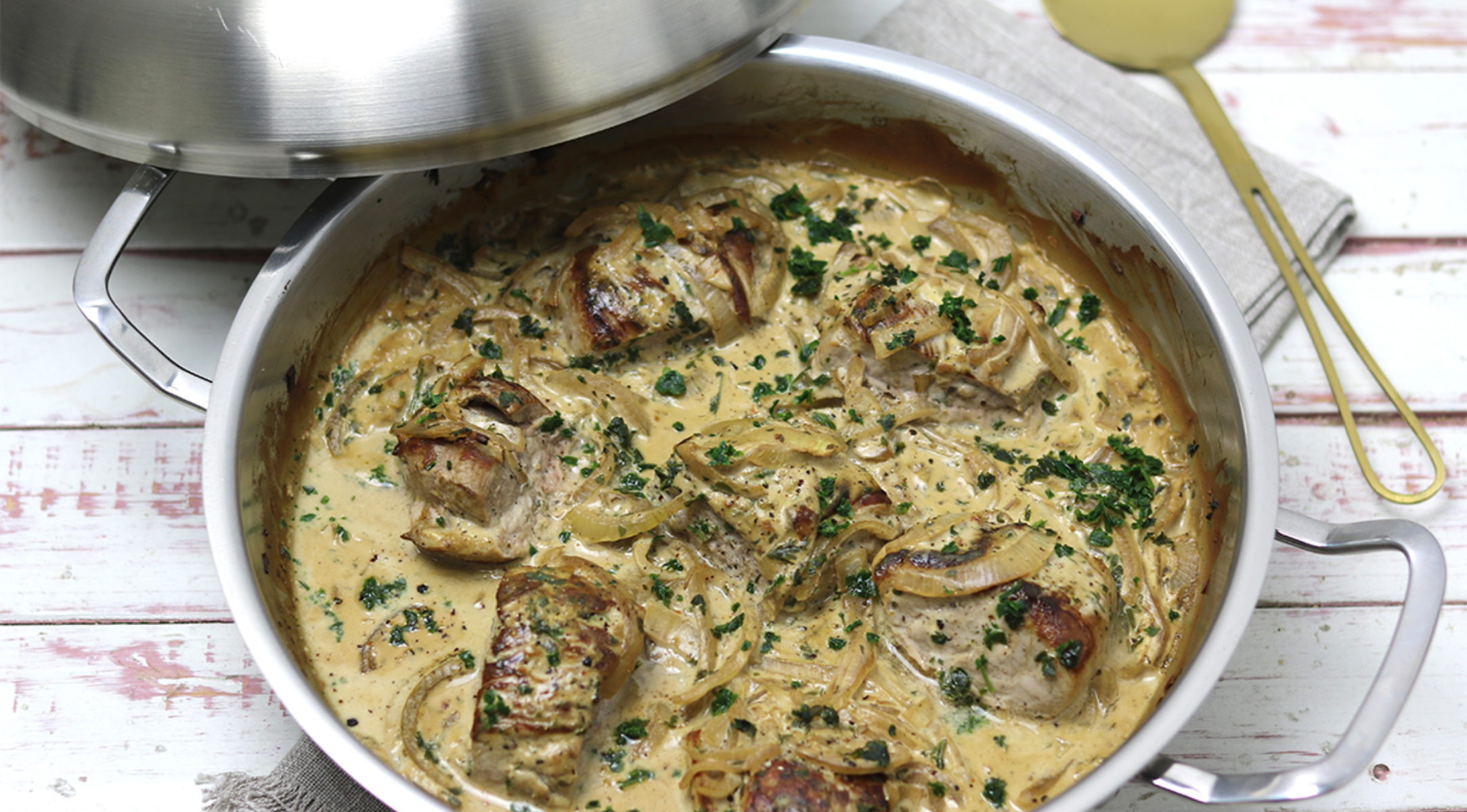 Fissler-Edelstahlpfanne mit Schweinemedaillons in Sahnesauce, Kräutern und Zwiebeln; teilweise mit Deckel bedeckt. Goldlöffel in der Nähe.
