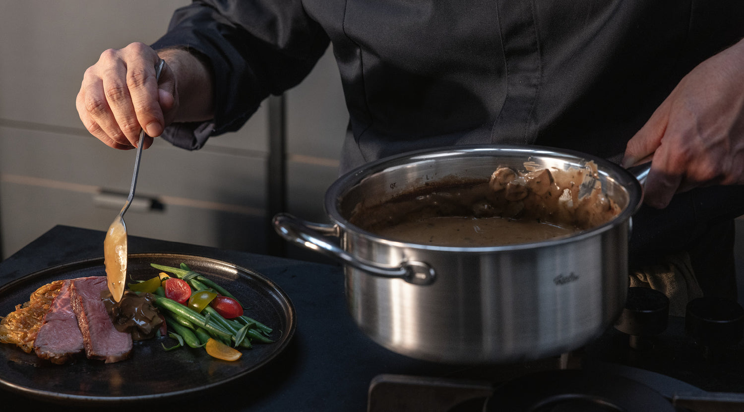 In einem Fissler-Edelstahltopf serviert man eine cremige Sauce über Fleisch, grüne Bohnen, Tomaten und Gemüse auf einem Teller.