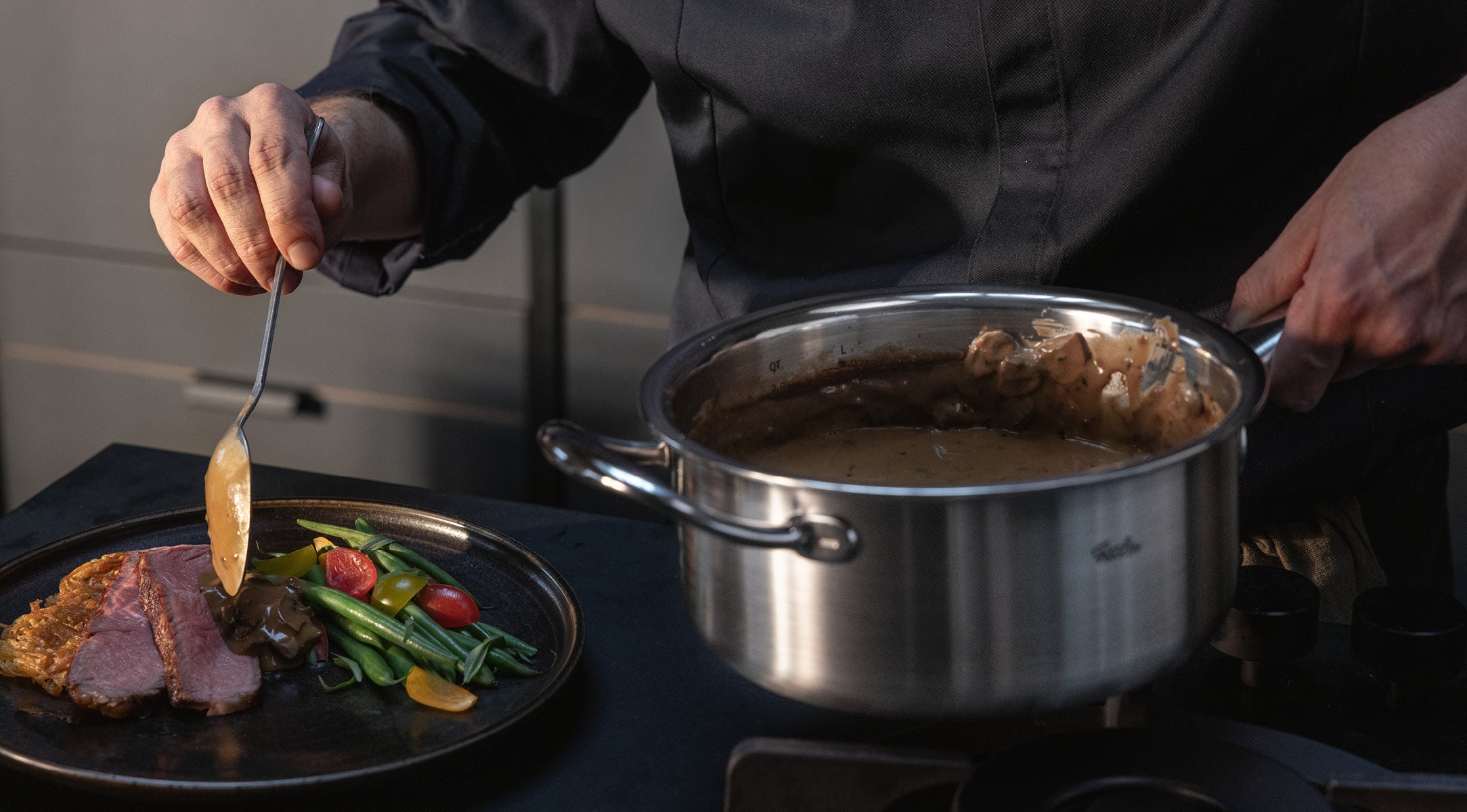 In einem Fissler-Edelstahltopf serviert man eine cremige Sauce über Fleisch, grüne Bohnen, Tomaten und Gemüse auf einem Teller.