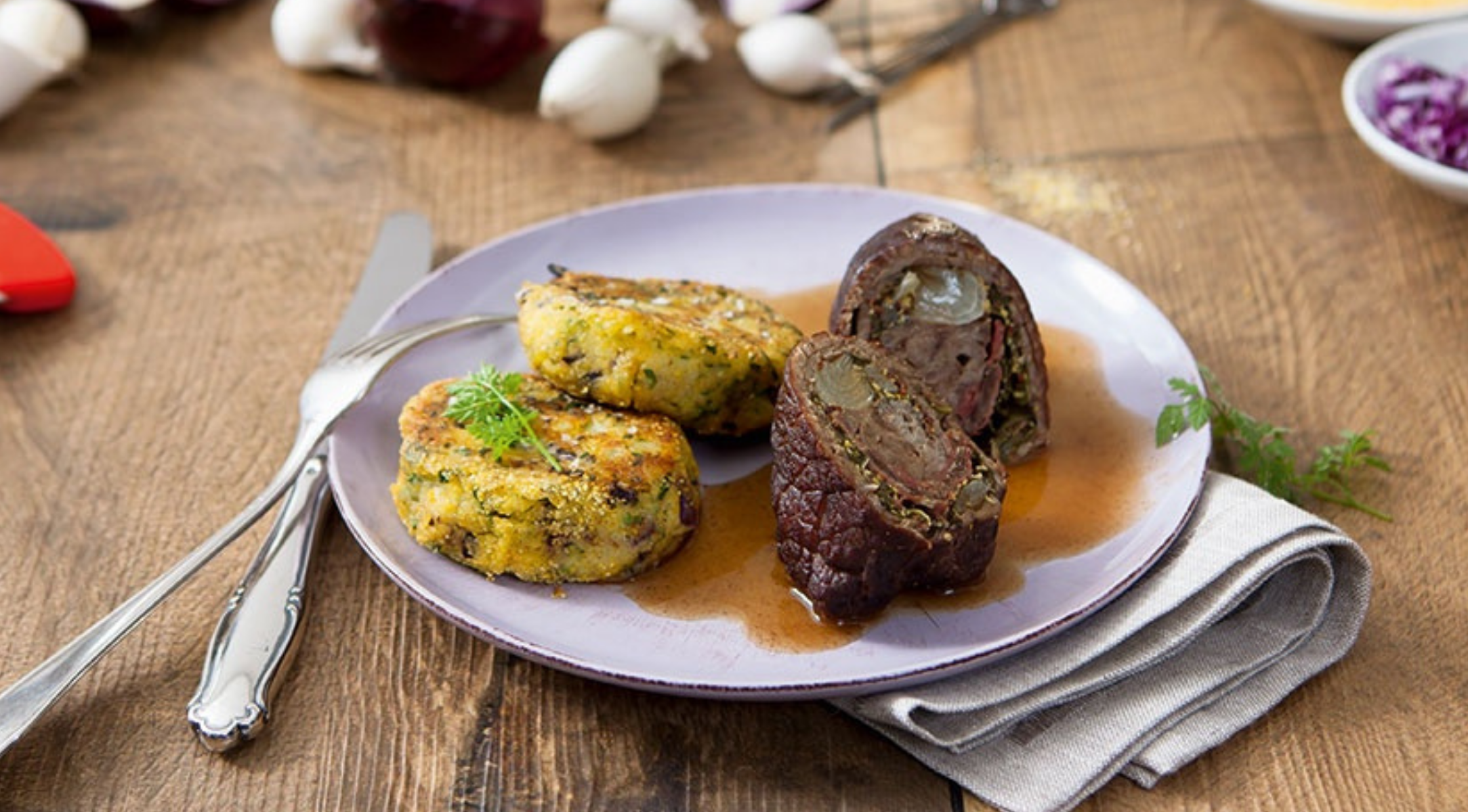 Eine klassische Rinderroulade mit Bratensoße und zwei Kartoffelknödeln, elegant auf einem Holztisch mit Fissler-Besteck angerichtet.