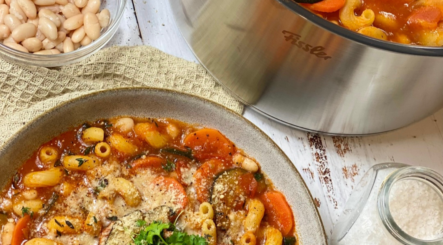 Eine Schüssel Suppe mit Kräutern und Käse neben einem Fissler-Edelstahltopf, Bohnen und Käse auf einer weißen Holzfläche.