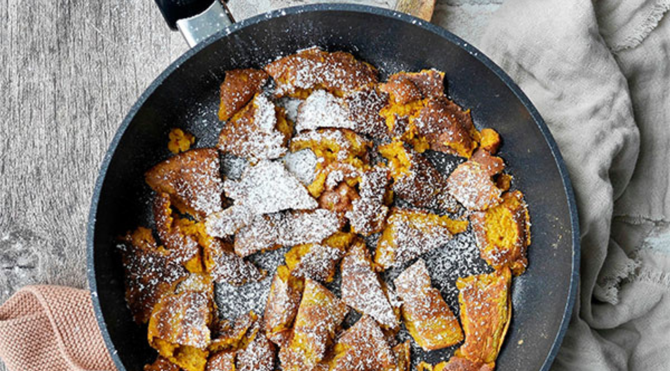 Eine hochwertige Fissler-Pfanne mit goldbraunen Pfannkuchenstücken, bestäubt mit Zucker, auf einer rustikalen Holzoberfläche neben einem beigen Tuch.