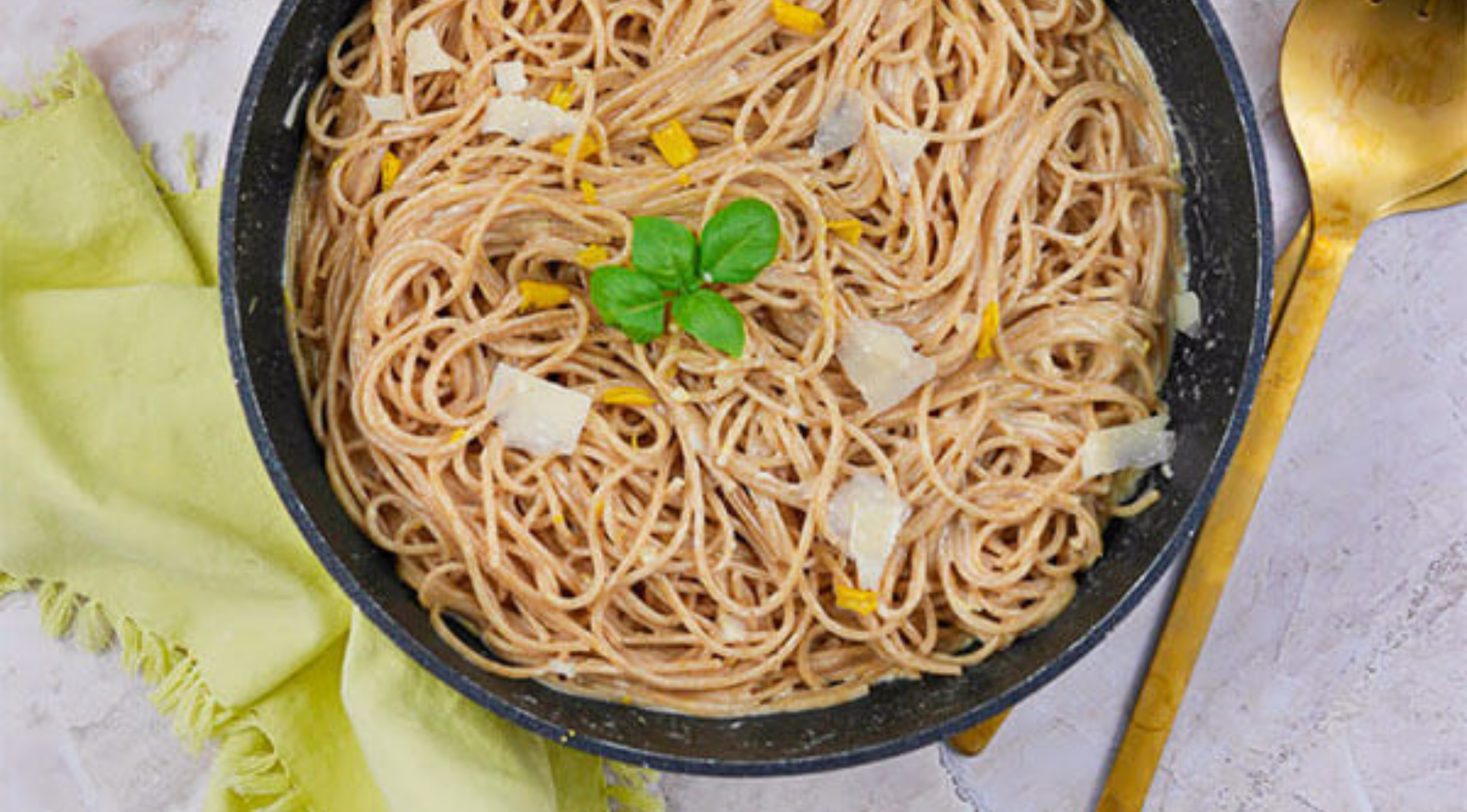 Frische Spaghetti mit geriebenem Käse und Basilikum in einer Schüssel, daneben ein goldener Löffel und eine Serviette auf Marmor, der mit Fissler-Kochgeschirr hergestellt wurde.
