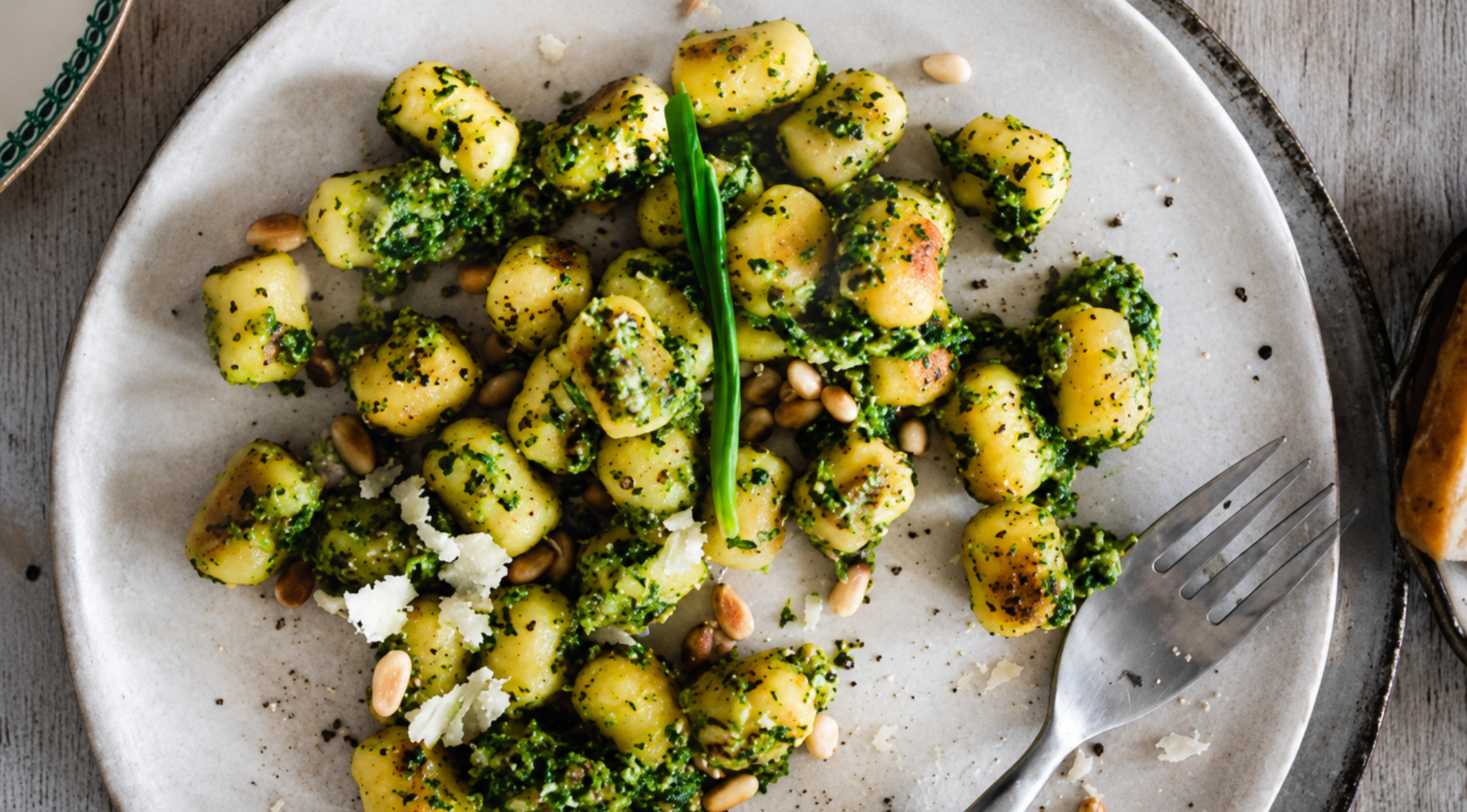 Eine wunderschön angerichtete Portion Gnocchi mit leuchtend grünem Pesto, garniert mit Pinienkernen und frisch geriebenem Käse, serviert auf einem eleganten Teller neben einer Edelstahlgabel – ein Beispiel für die zeitlose Qualität und das Design, die das Kochgeschirr von Fissler auszeichnen.