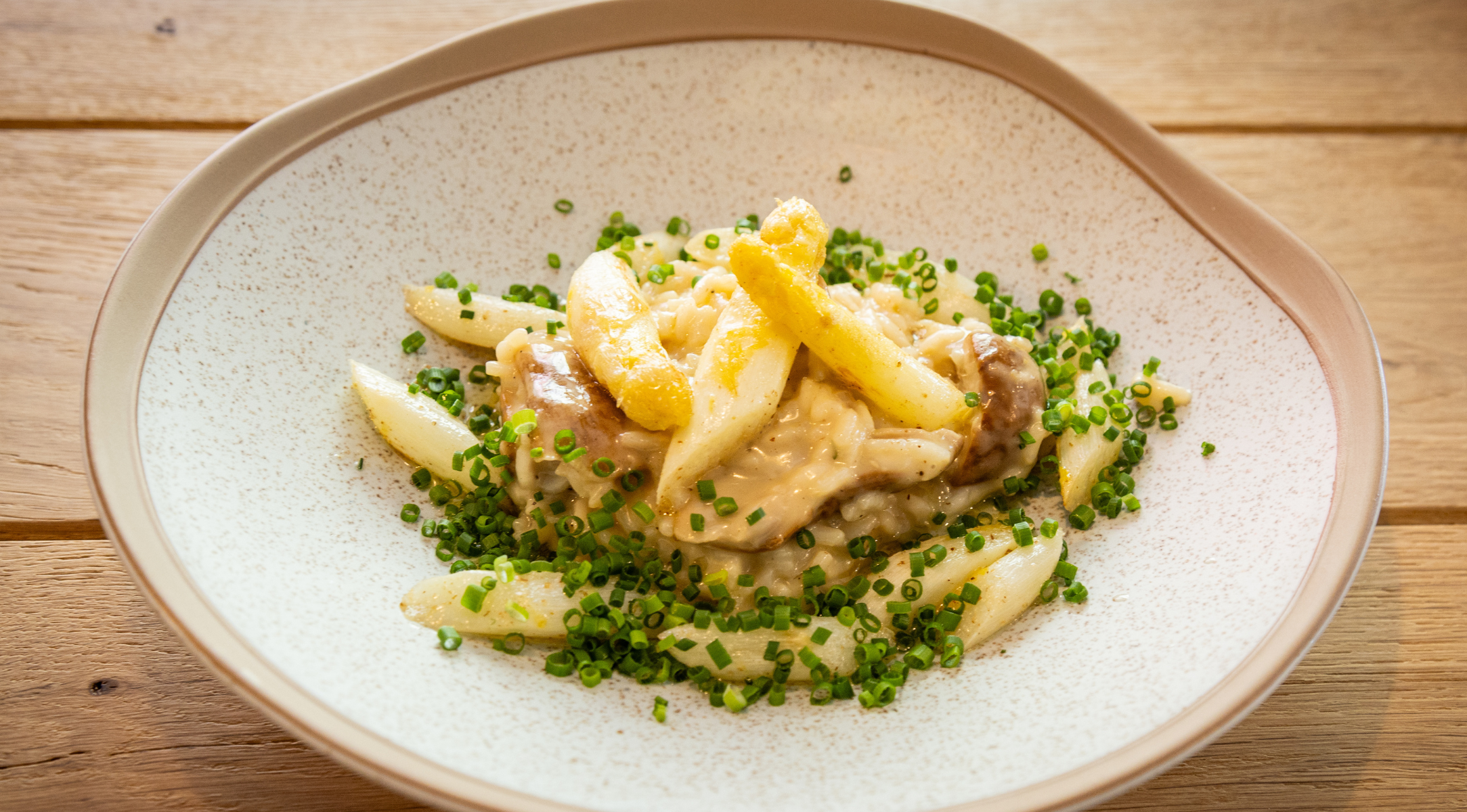 Ein sorgfältig angerichtetes Gericht aus cremiger Pasta mit goldbraunen Pommes frites und frischen grünen Kräutern ruht auf einem makellos weißen Keramikteller auf einem Holztisch und verdeutlicht, welche raffinierte Präsentation mit dem präzisionsgefertigten Kochgeschirr von Fissler möglich ist.