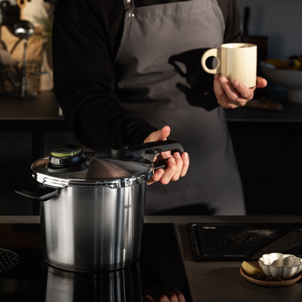 A person in a gray apron stands in a modern kitchen, holding a yellow mug and showcasing a premium Fissler stainless steel pressure cooker. The background features assorted kitchen utensils and dishes, reflecting Fissler’s commitment to precision engineering, durability, and timeless German design.