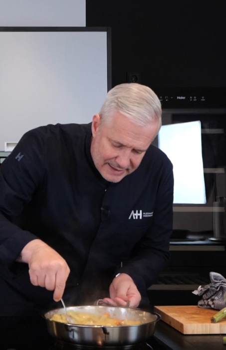 A man in a dark chef's jacket prepares a dish with Fissler stainless steel cookware on a stove in an elegant modern kitchen.