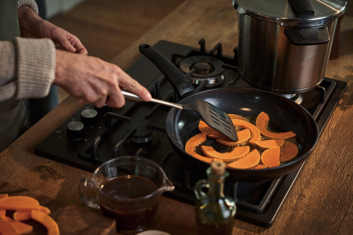 A person prepares sliced pumpkin in a Fissler frying pan on a gas stove, using a precision-engineered spatula. On the premium wooden counter, a durable Fissler stainless steel pot, a glass measuring cup with liquid, and an elegantly designed bottle of oil highlight the timeless quality and German craftsmanship of Fissler cookware.