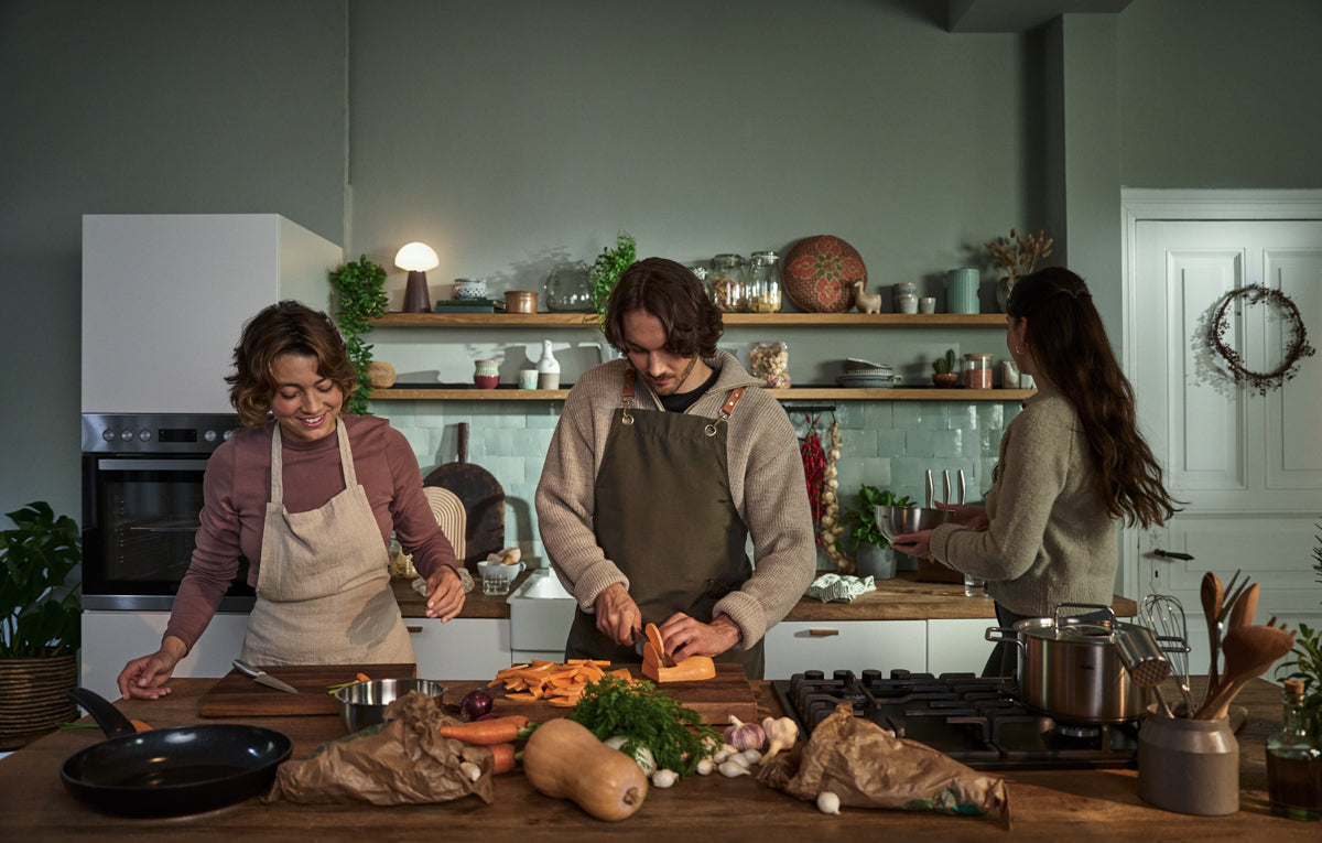 Three people in a kitchen prepare food together, showcasing Fissler’s stainless steel cookware. One person slices sweet potatoes on a wooden counter while another works nearby using Fissler pots and pans, and a third stands at the sink. A variety of fresh vegetables and premium Fissler kitchen accessories are arranged on the countertop, reflecting precision engineering and timeless German design.