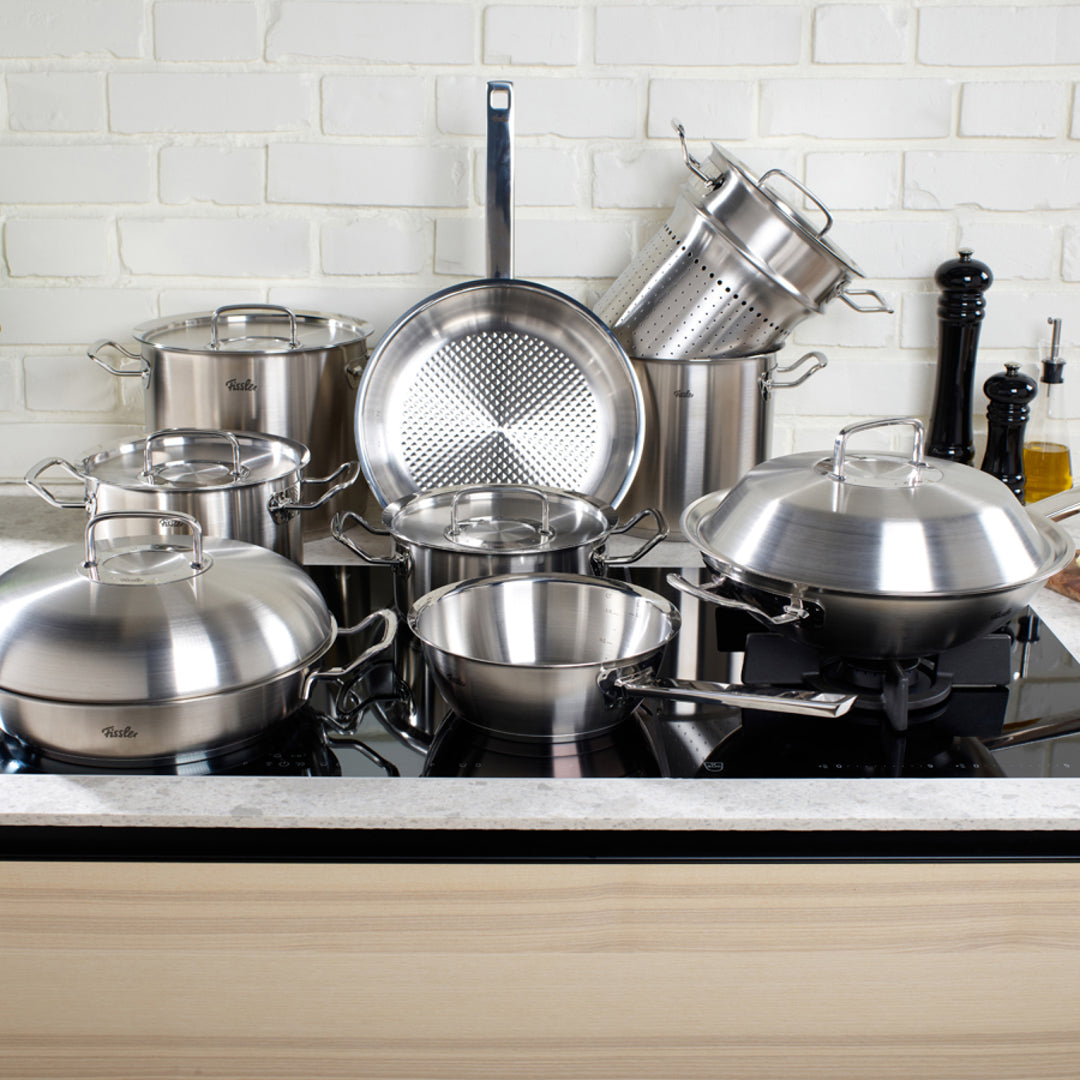 Töpfe, Pfannen, Siebe und Deckel aus rostfreiem Stahl von Fissler auf einem Küchentisch mit weißem Backsteinsockel; Qualität aus deutscher Produktion.