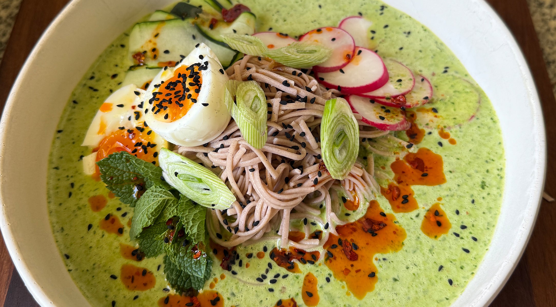 Eine wunderschön angerichtete Schüssel Soba-Nudeln mit Radieschenscheiben, einem weichgekochten Ei, knackiger Gurke, Frühlingszwiebeln, frischer Minze, schwarzem Sesam und Chiliöl in einer cremigen grünen Soße – präsentiert, um die Präzision und Kunstfertigkeit zu verdeutlichen, die Fissler-Kochgeschirr in Ihre Küche bringt.