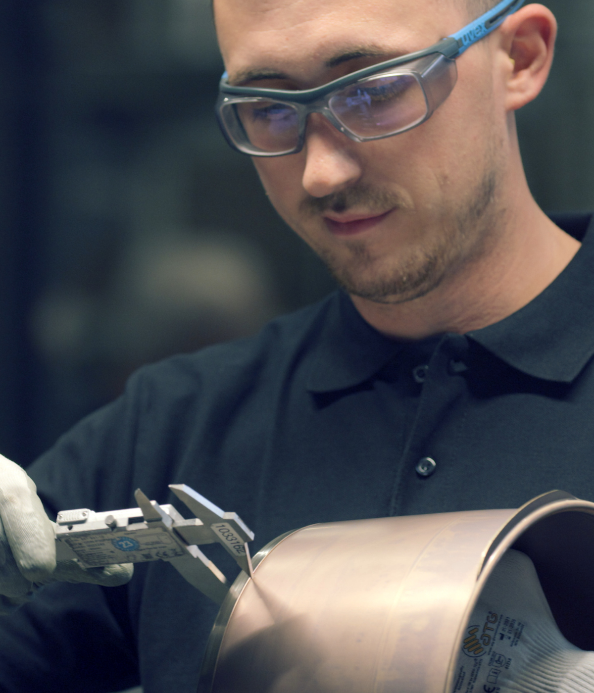 Ein Handwerker mit Schutzbrille und Handschuhen misst ein Topfteil aus rostfreiem Stahl aus und stellt die deutsche Präzisionstechnik von Fissler unter Beweis.